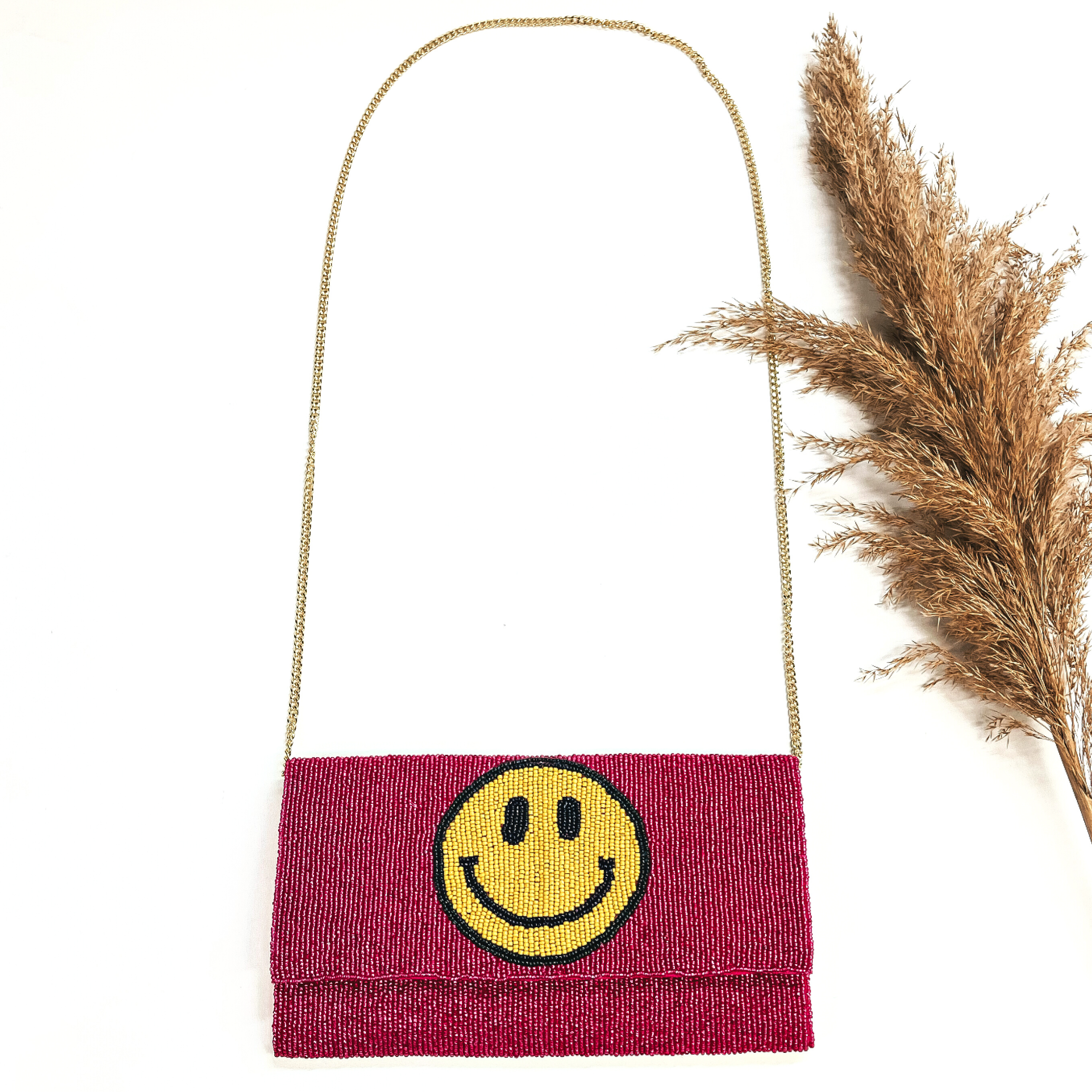 This is a seedbeaded clutch purse with a gold tone chain. This bag is in fuchsia with  a yellow happy face.This bag  is taken laying on a white background with a brown plant in the side as decor.