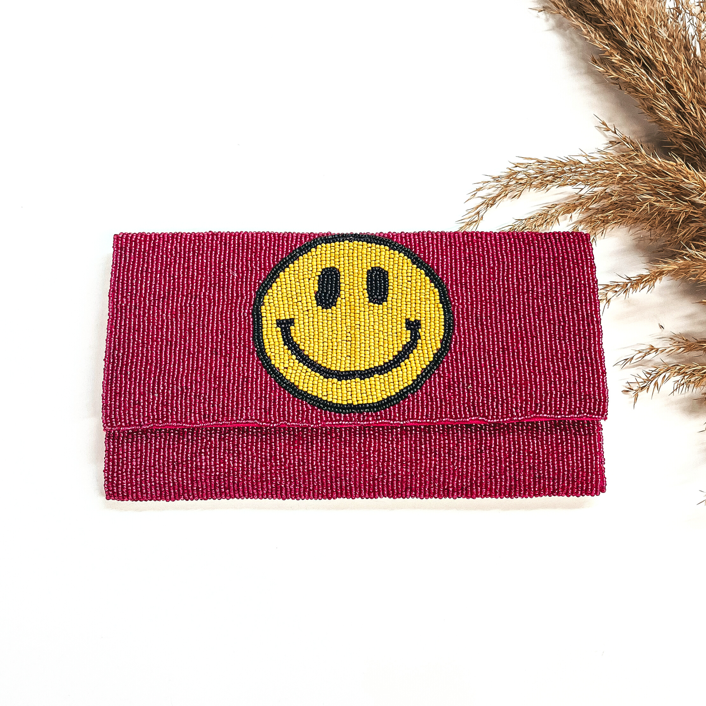 This is a seedbeaded clutch purse in fuchsia with a yellow happy face.This bag  is taken laying on a white background with a brown plant in the side as decor.