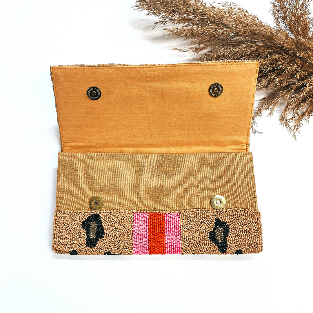 This is the inside of the seedbeaded clutch purse in brown and leopard print in  black and gold with  three stripes in the middle, two hot pink and one orange line in the middle. This bag  is taken laying on a white background with a brown plant in the side as decor.