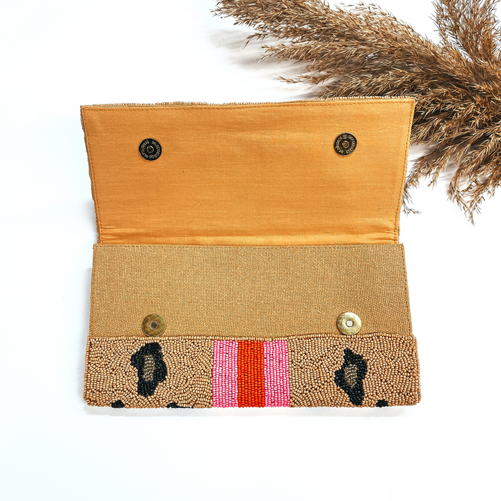 This is the inside of the seedbeaded clutch purse in brown and leopard print in  black and gold with  three stripes in the middle, two hot pink and one orange line in the middle. This bag  is taken laying on a white background with a brown plant in the side as decor.