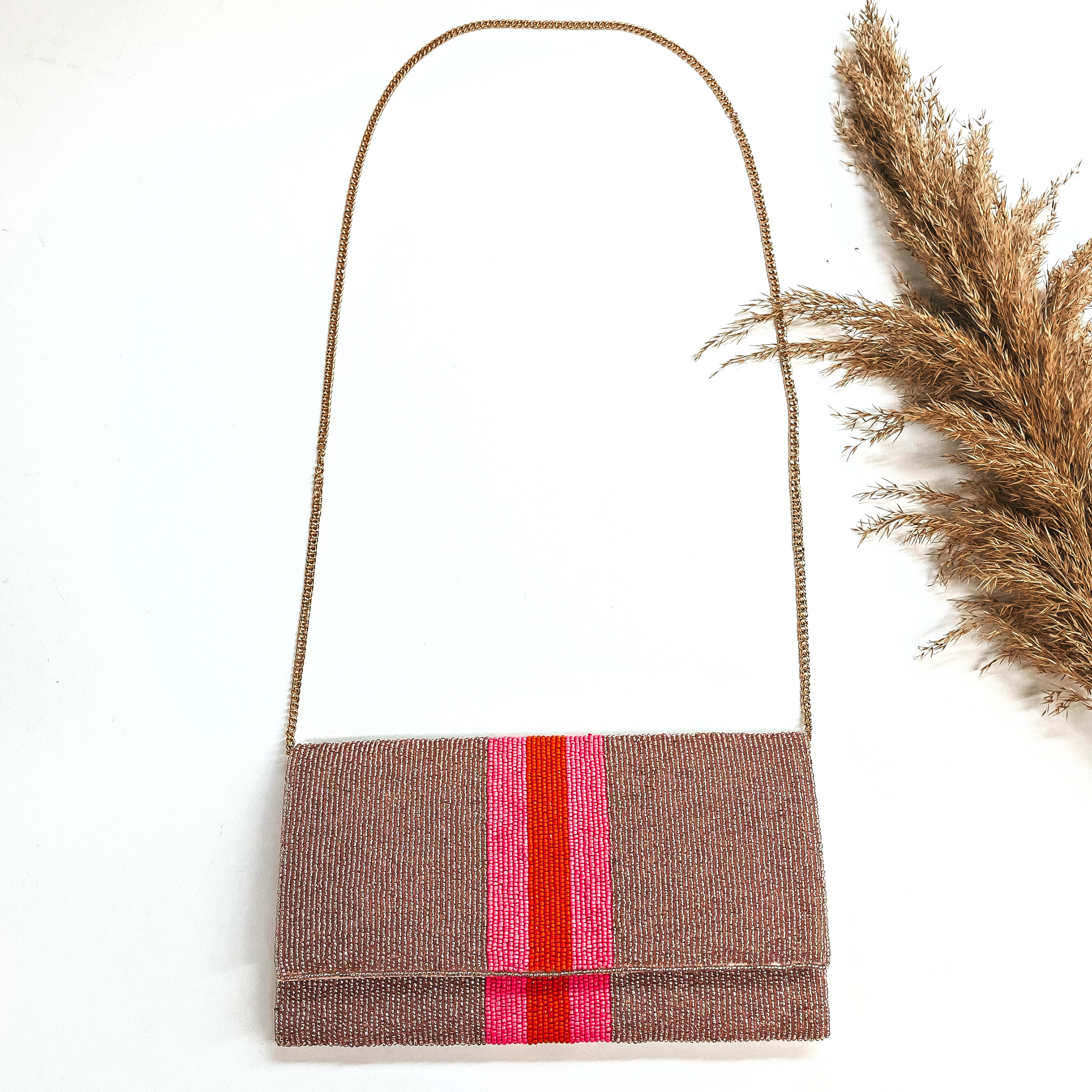 This is a seedbeaded clutch purse with a gold tone chain. This bag is in rose gold with  three stripes in the middle, two hot pink and one orange line in the middle. This bag  is taken laying on a white background with a brown plant in the side as decor.