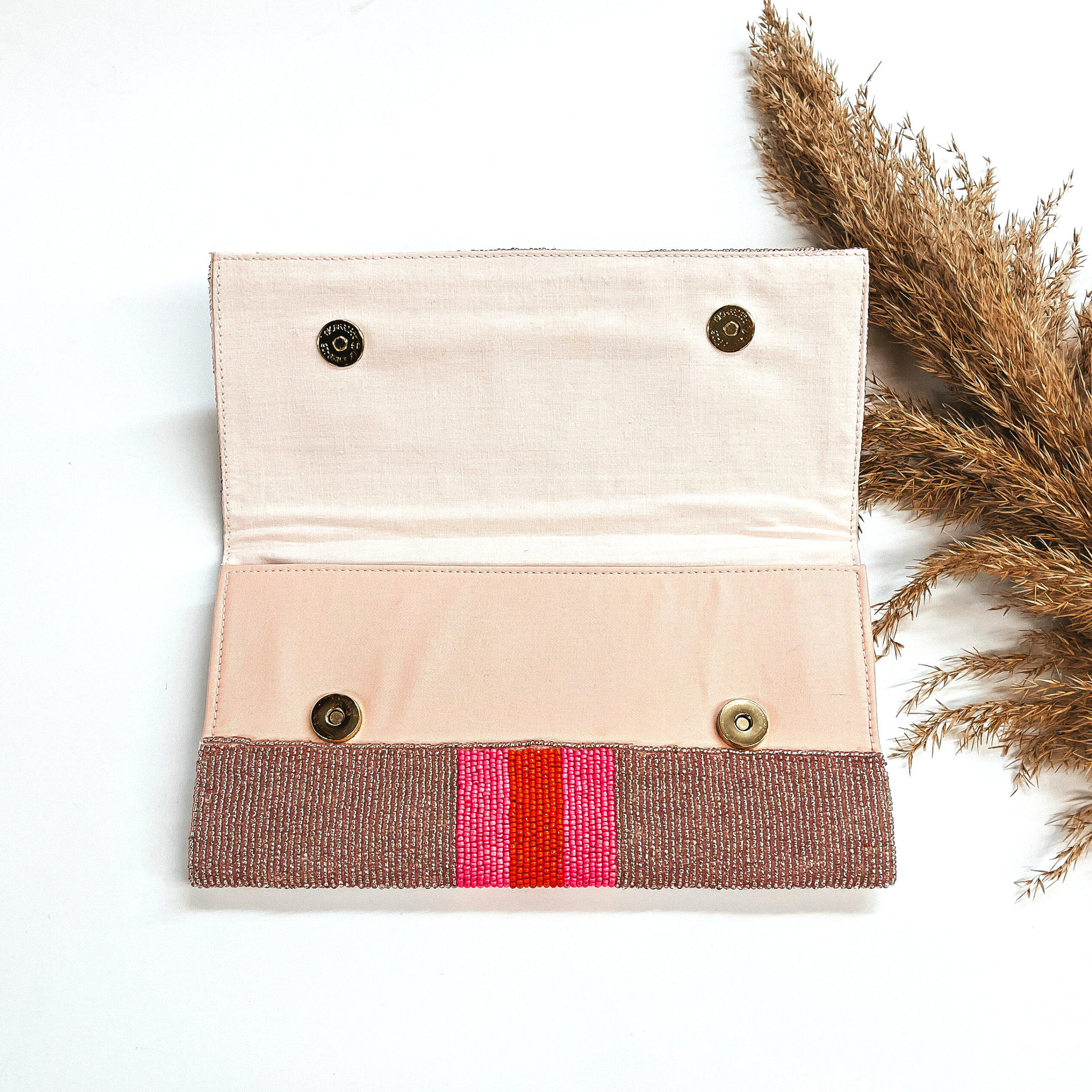 This is the inside of the seedbeaded clutch purse in rose gold with  three stripes in the middle, two hot pink and one orange line in the middle. This bag  is taken laying on a white background with a brown plant in the side as decor.