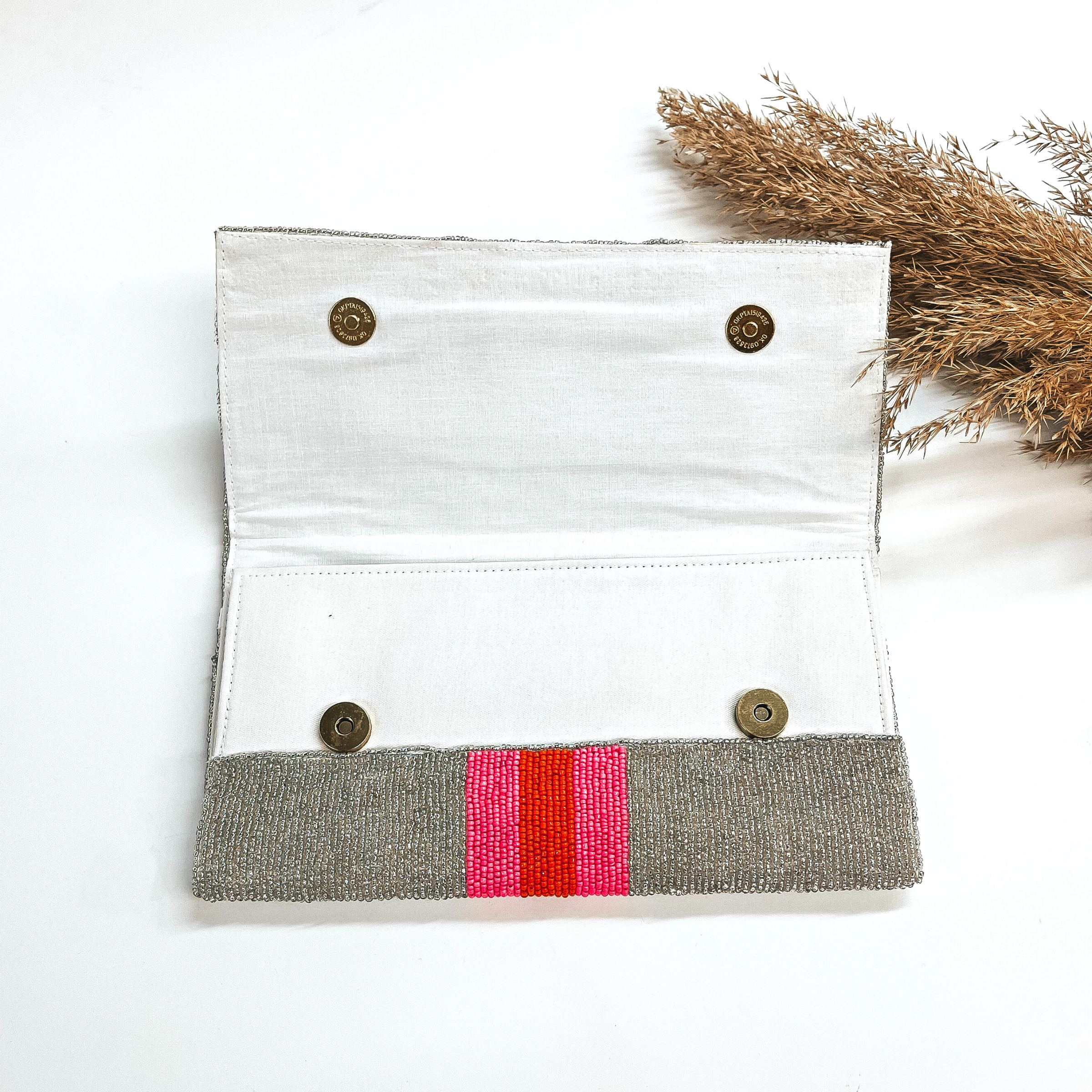 This is the inside of the seedbeaded clutch purse in silver with  three stripes in the middle, two hot pink and one orange line in the middle. This bag  is taken laying on a white background with a brown plant in the side as decor.