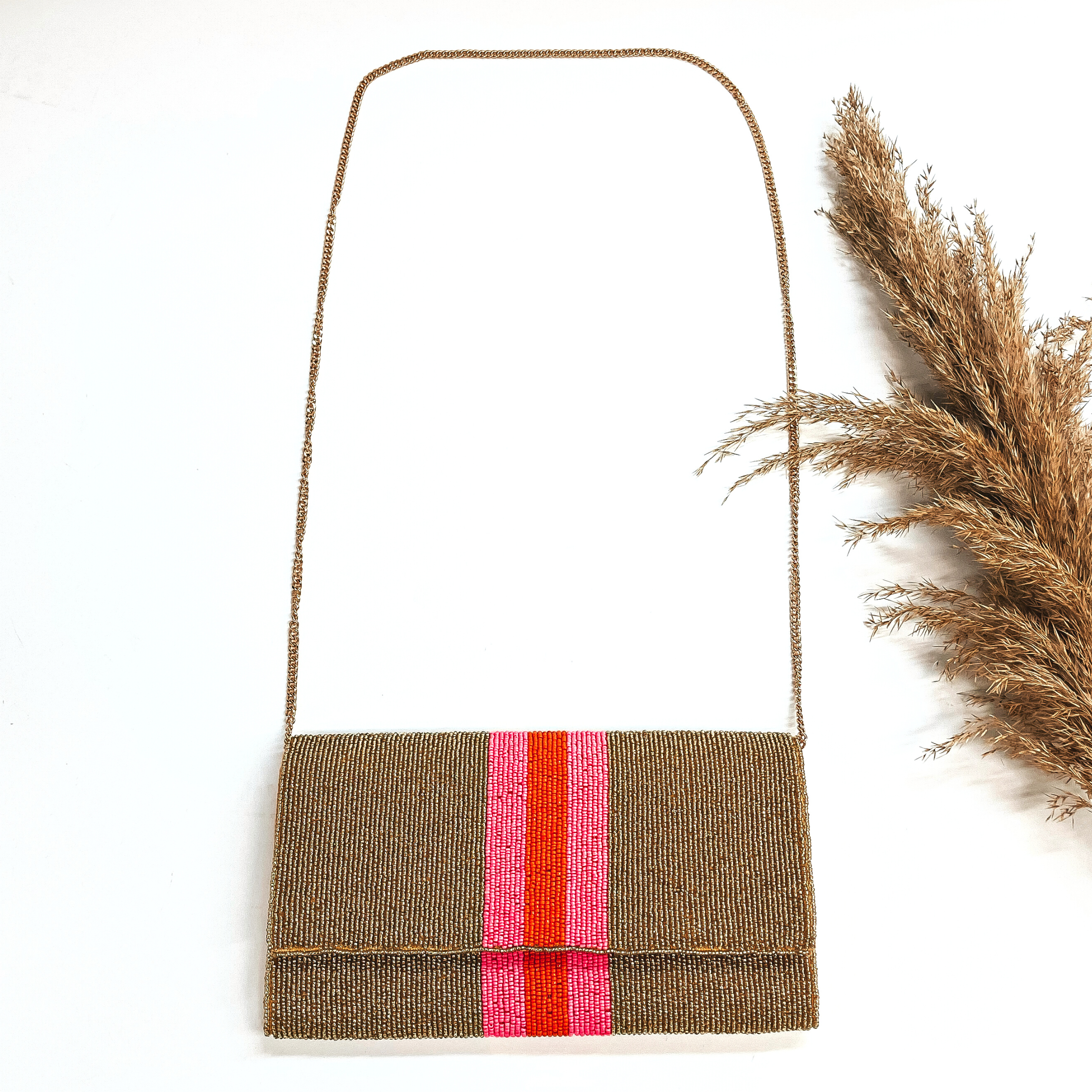 This is a seedbeaded clutch purse with a gold tone chain. This bag is in  gold with  three stripes in the middle, two hot pink and one orange line in the middle. This bag  is taken laying on a white background with a brown plant in the side as decor.