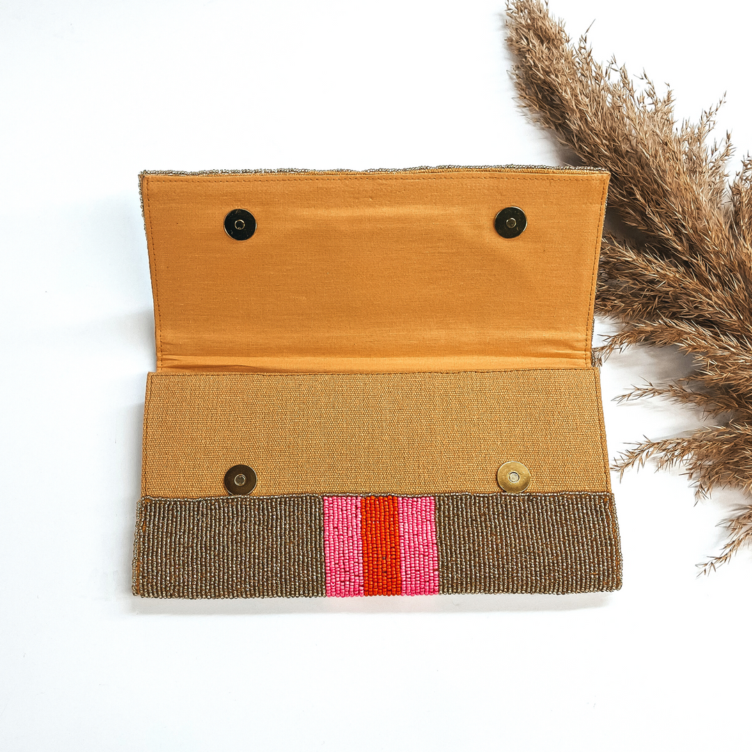 This is the inside of the seedbeaded clutch purse in gold with  three stripes in the middle, two hot pink and one orange line in the middle. This bag  is taken laying on a white background with a brown plant in the side as decor.