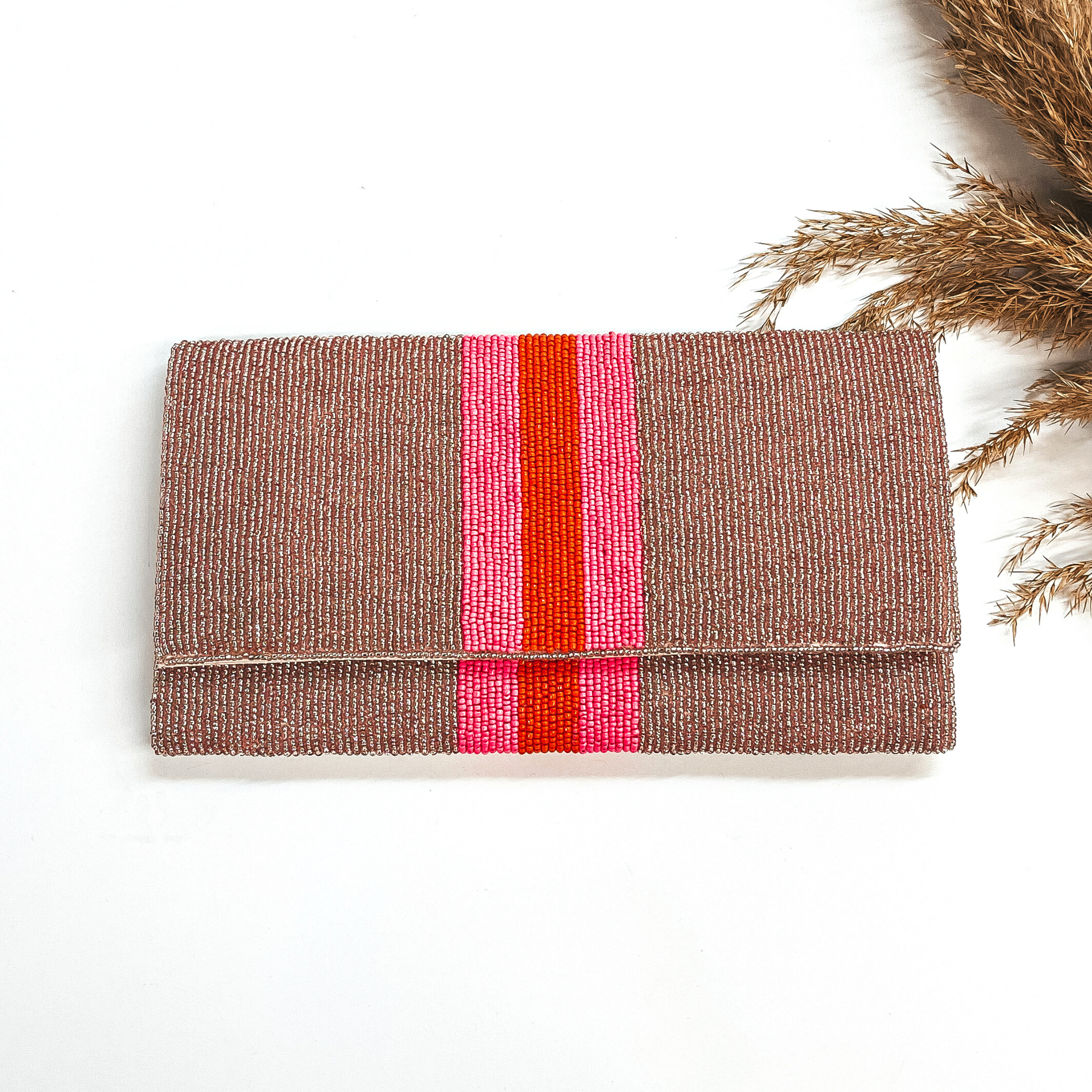 This is a seedbeaded clutch purse in rose gold with  three stripes in the middle, two hot pink and one orange line in the middle. This bag  is taken laying on a white background with a brown plant in the side as decor.