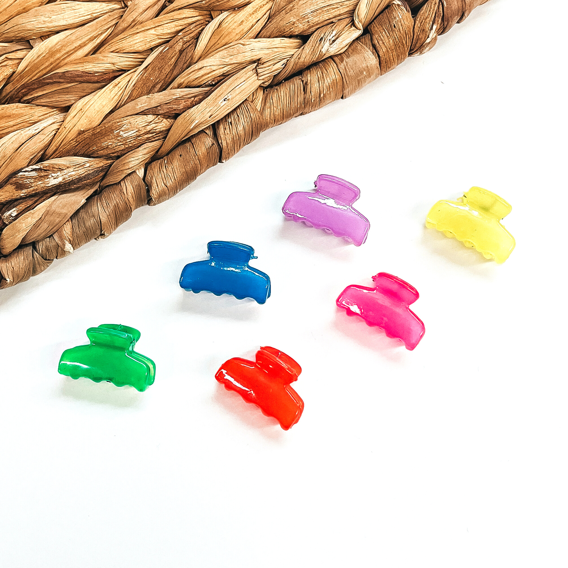 There are six small colorful hair clips on a white background and a brown woven slate  on the side as decor. From left to right; neon green, neon orange, blue, neon pink,  purple, and neon yellow. 