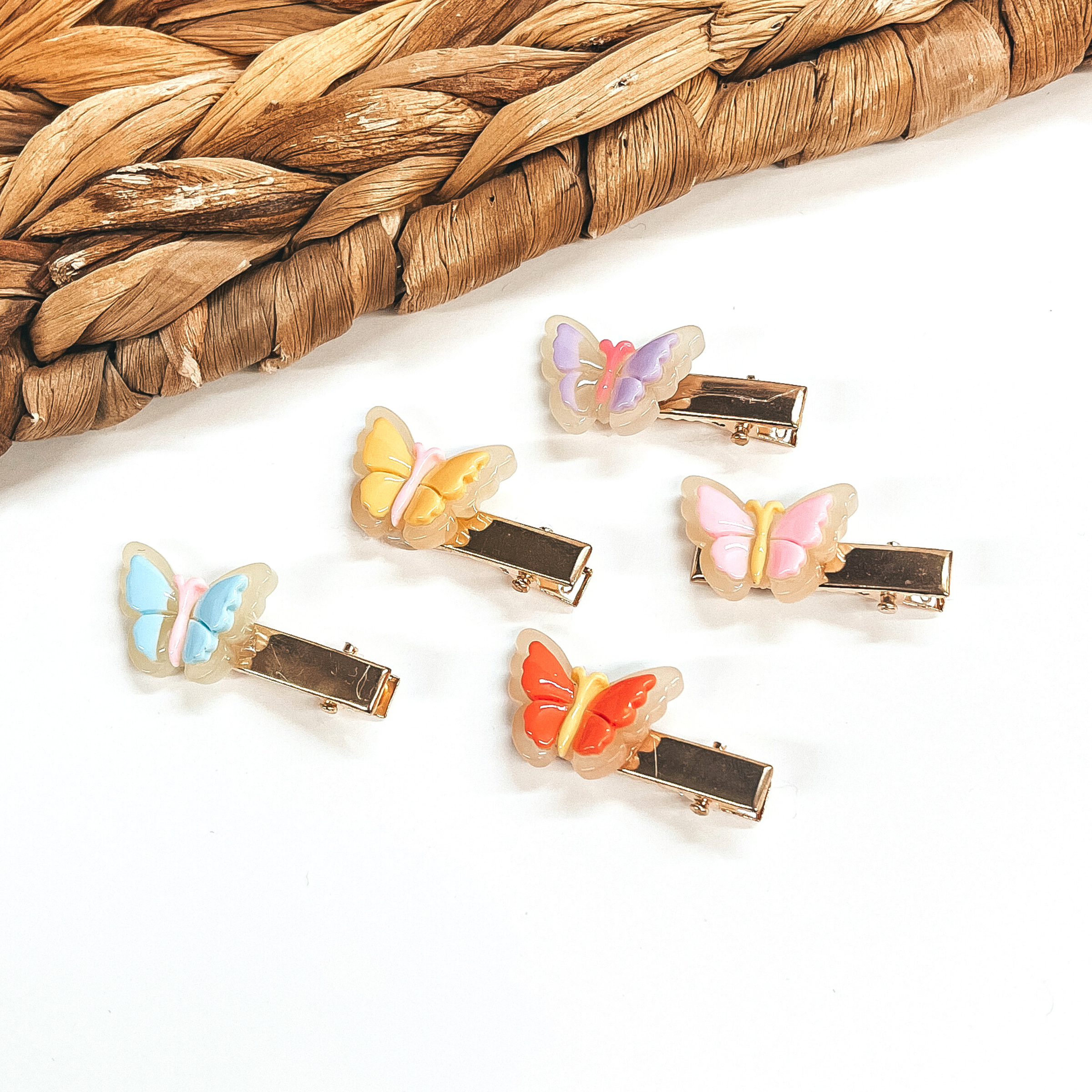 There are five small gold clips with a 3D butterfly charm on them. From left to right;  light blue with light pink body, orange with yellow body, yellow with light pink  body, light pink with yellow body, and light purple with pink body.  These clips are laying on a white background  with a brown woven slate in the back as decor.