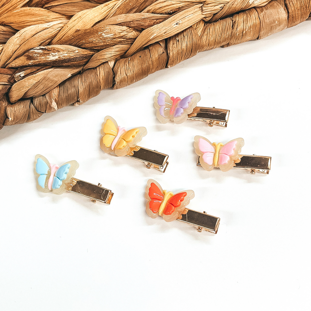 There are five small gold clips with a 3D butterfly charm on them. From left to right;  light blue with light pink body, orange with yellow body, yellow with light pink  body, light pink with yellow body, and light purple with pink body.  These clips are laying on a white background  with a brown woven slate in the back as decor.