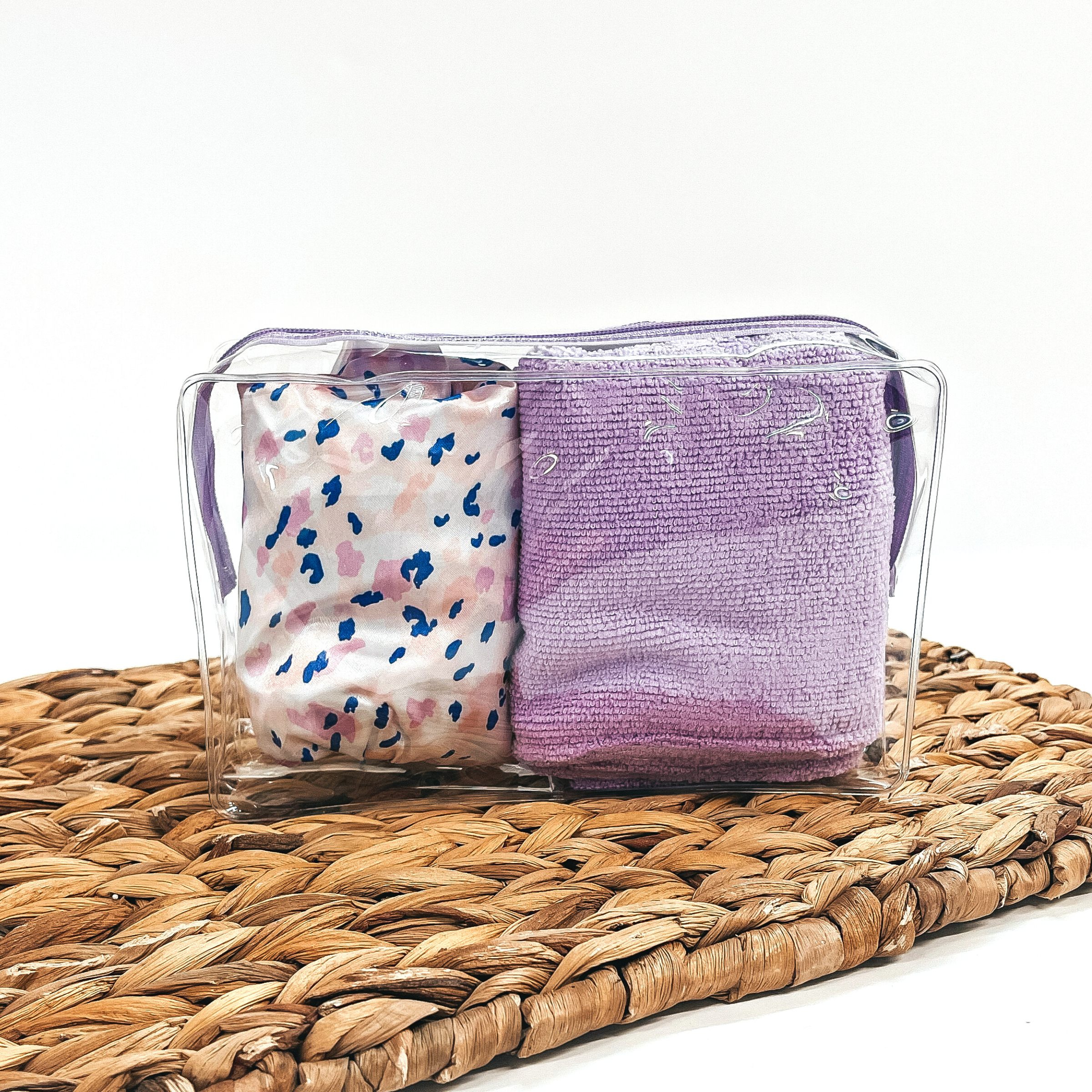 This is a clear bag with a purple lavender zipper, inside the bag there is a pink and  purple leopard shower cap and a purple hair turban. This bag is laying on a brown  woven slate and on a white background.