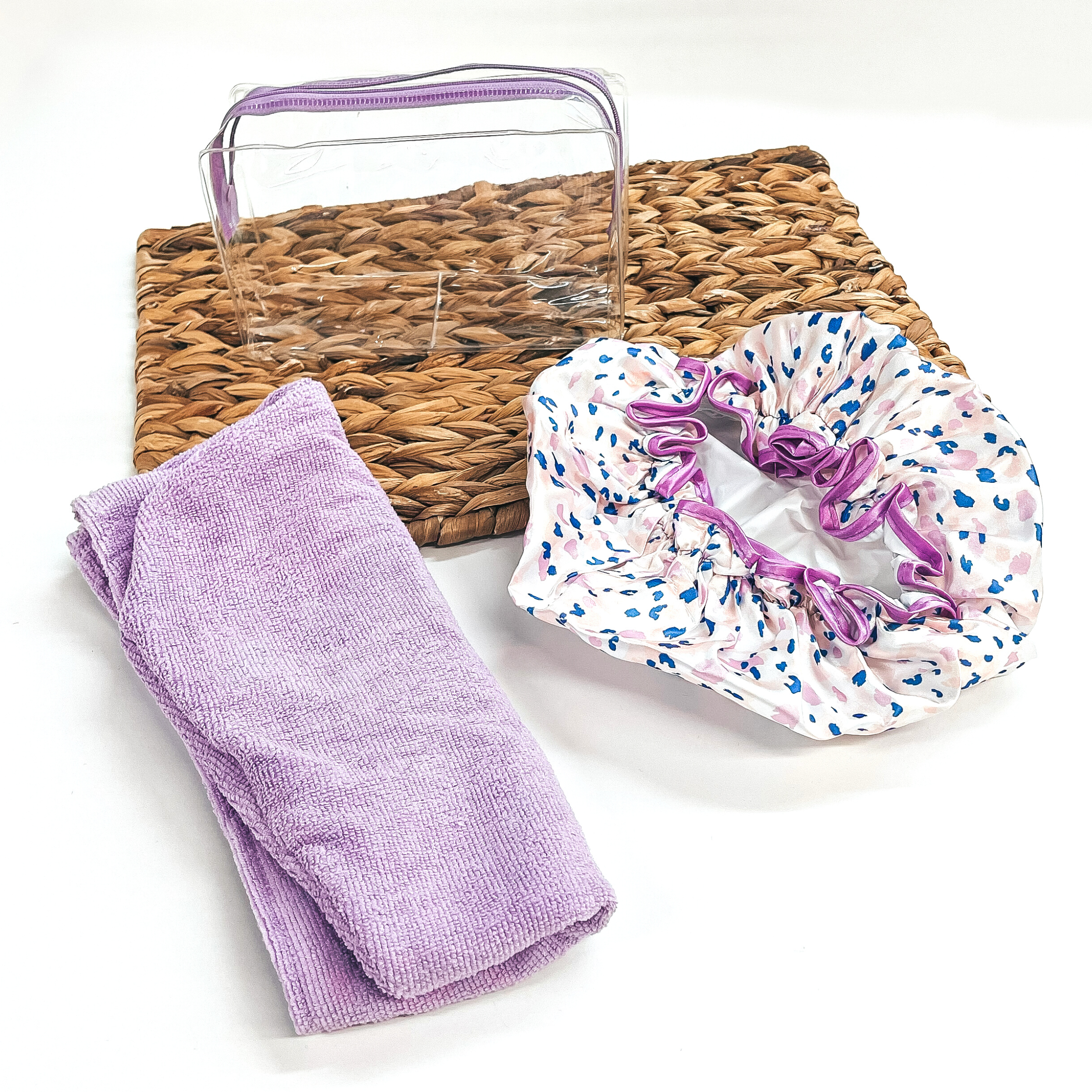 There is a clear bag with a purple lavender zipper on top of a brown woven slate.  There is a purple hair turban and a pink and purple leopard print shower cap laying  partly on a brown woven slate and on a white background.
