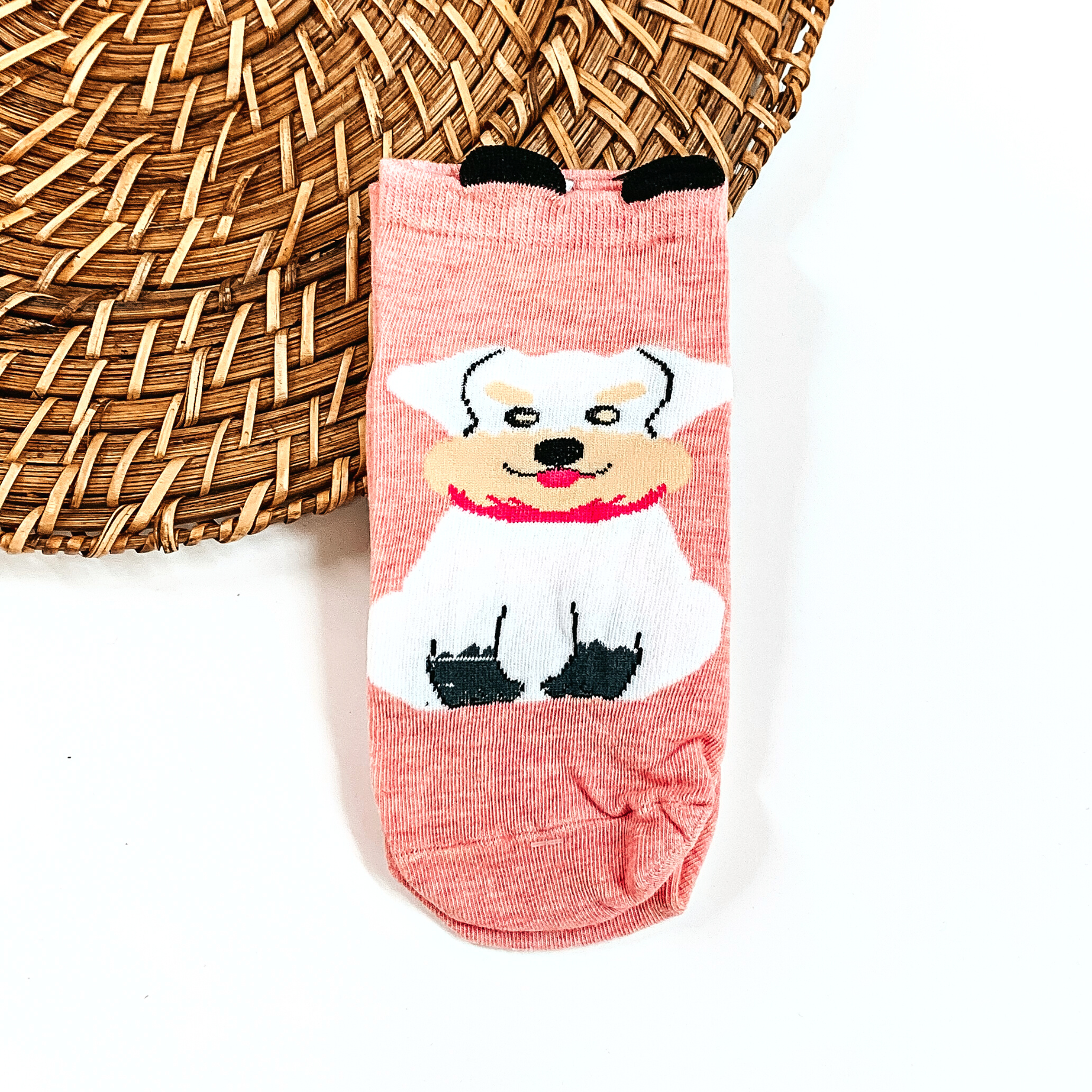 This is a light pink pair of socks with a white Schnauzer dog and a pink scarf.  They have small black 'ears' on the top. This pair of socks are taken on a brown  woven plate and on a white background.