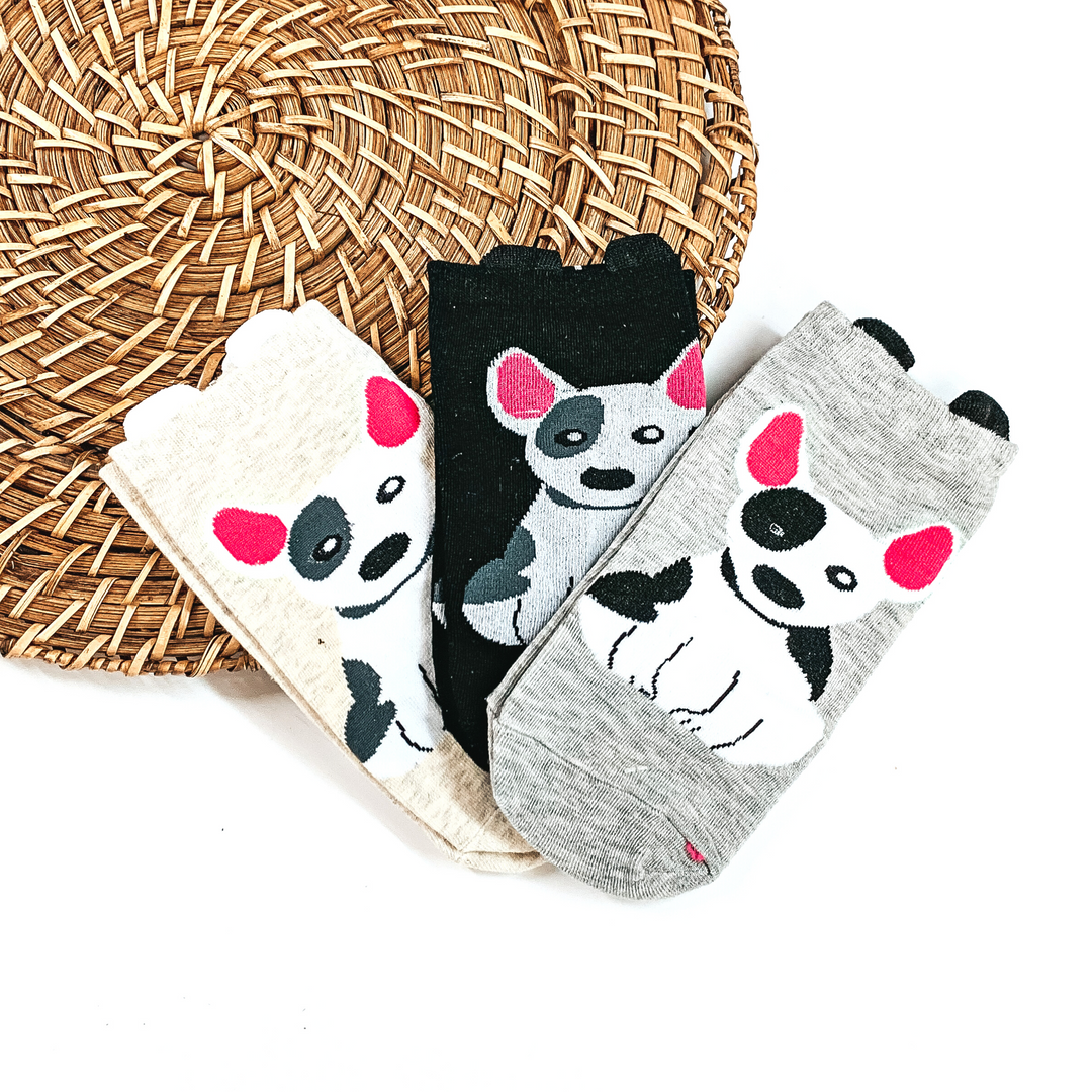 There are three pairs of socks with a Bull Terrier dog in the center in three colors. From left to tight; beige with a grey/white dog, black with a white/grey dog, and  then a grey pair of socks with black/white dog. These three pairs of socks are taken on  a brown woven slate and on a white background.
