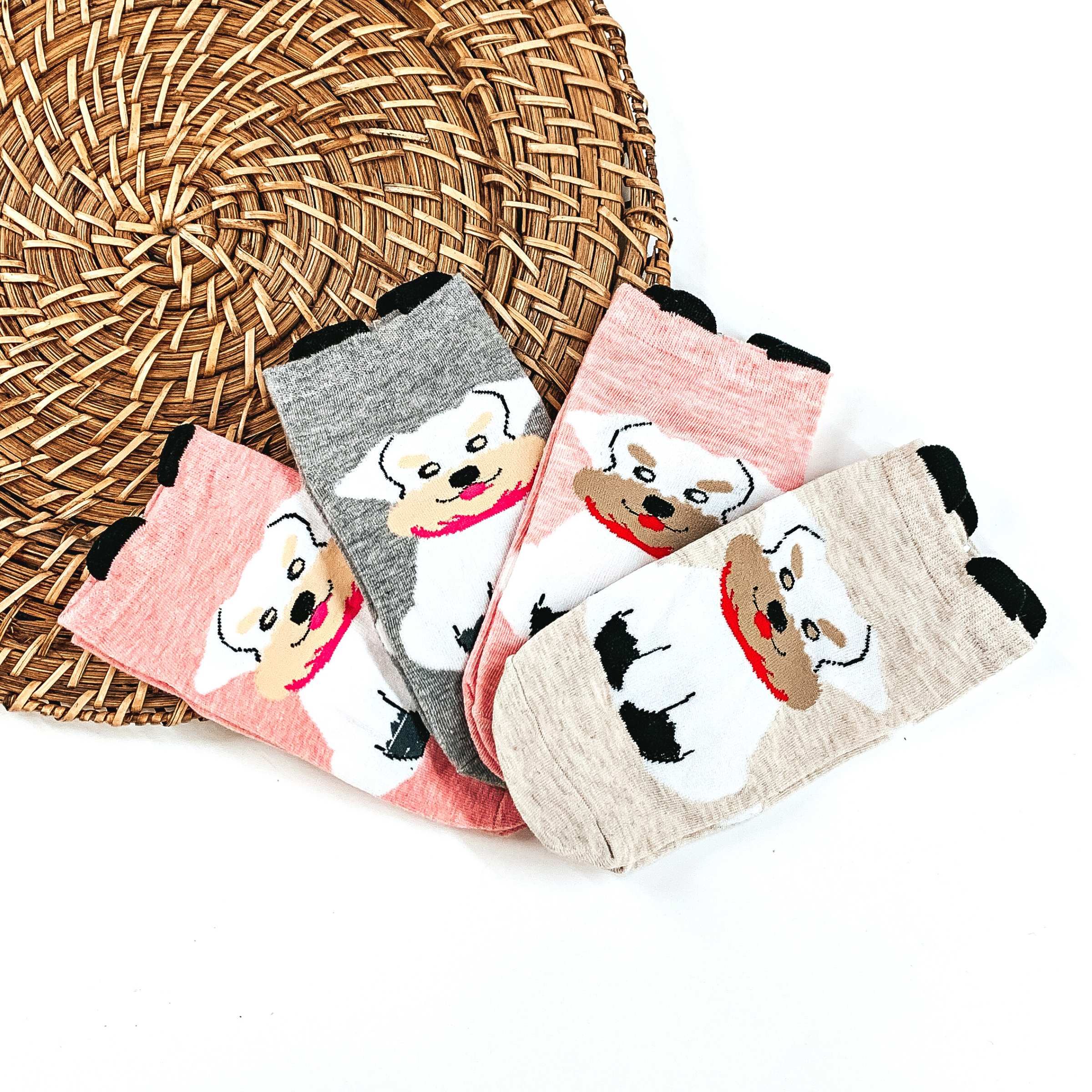 There are four pairs of socks in different colors with a Schnauzer dog in the center  and small black 'ears' on the top.  From left to right; light pink with white Schnauzer and pink scarf, grey with white  Schnauzer and pink scarf, light pink with white Schnazuer and red scarf, and the last one is beige with a white Schnauzer with a red scarf. These pairs of socks are taken  laying on a brown woven plate and on a white background.