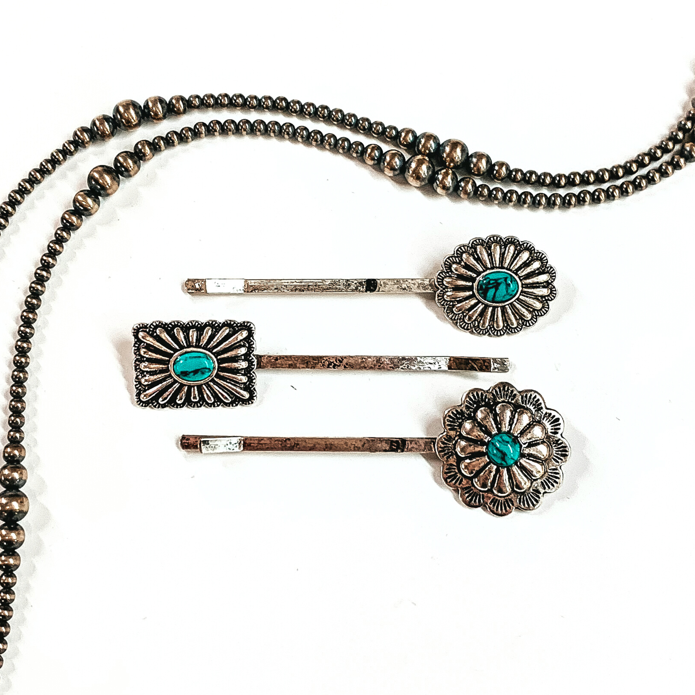 There are three silver bobby pins with conchos and a small faux turquoise stones  in the center of the concho. From top to bottom; an oval concho, rectangle/square  concho, and then a flower concho. These bobby pins are taken on a white background  with silver navajo pearls around as decor.
