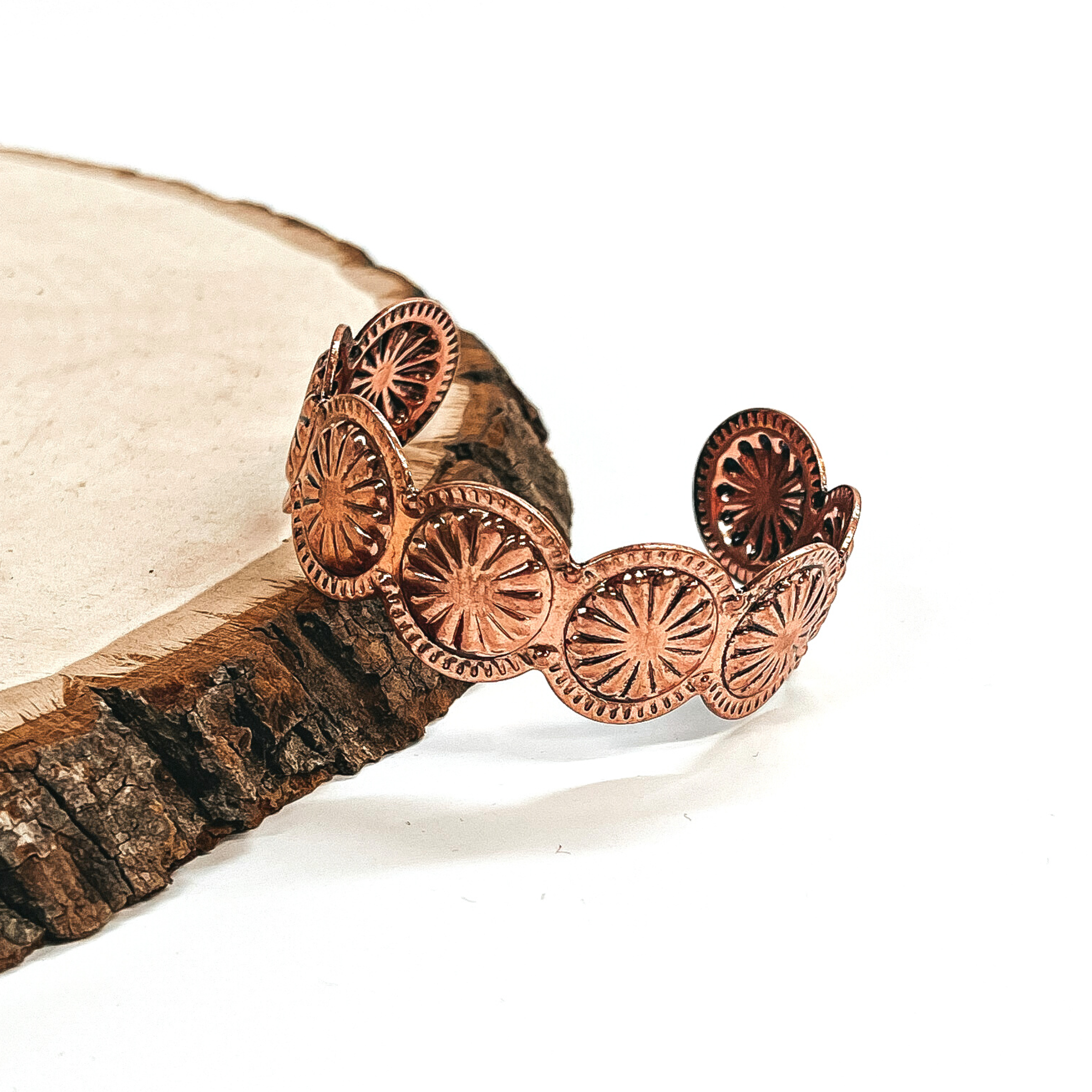 This is a copper concho stamped cuff bracelet leaning against a slab of wood and white  background.
