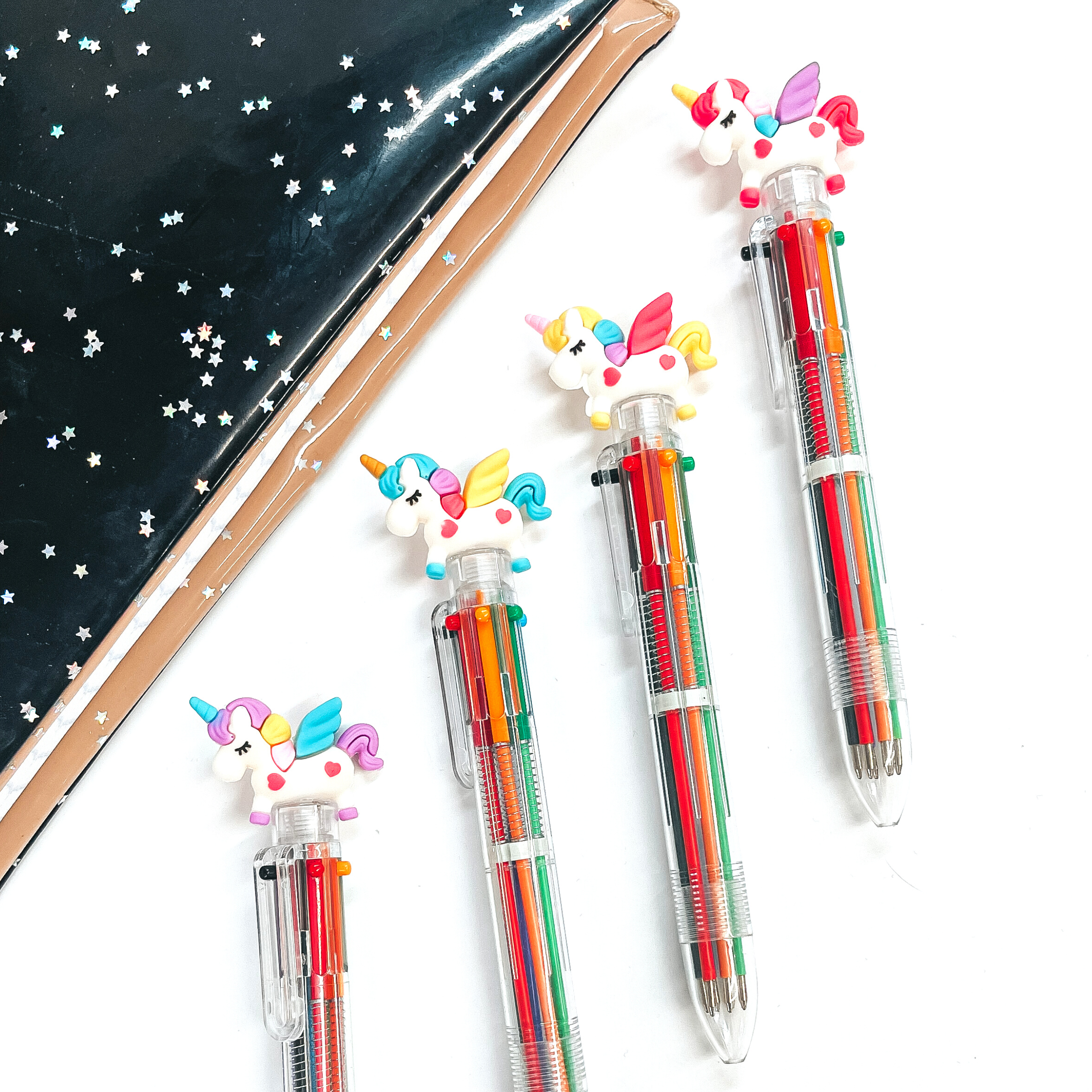 There are four shuttle pens with different colored unicorn toppers. All pens  have the same number and colored inks. From left to right; purle hair unicorn,  turquoise hair unicorn, yellow hair unircorn, and pink hair unicorn. All of these  pens are laying on a white background with a black shiny notebook and shiny stars.