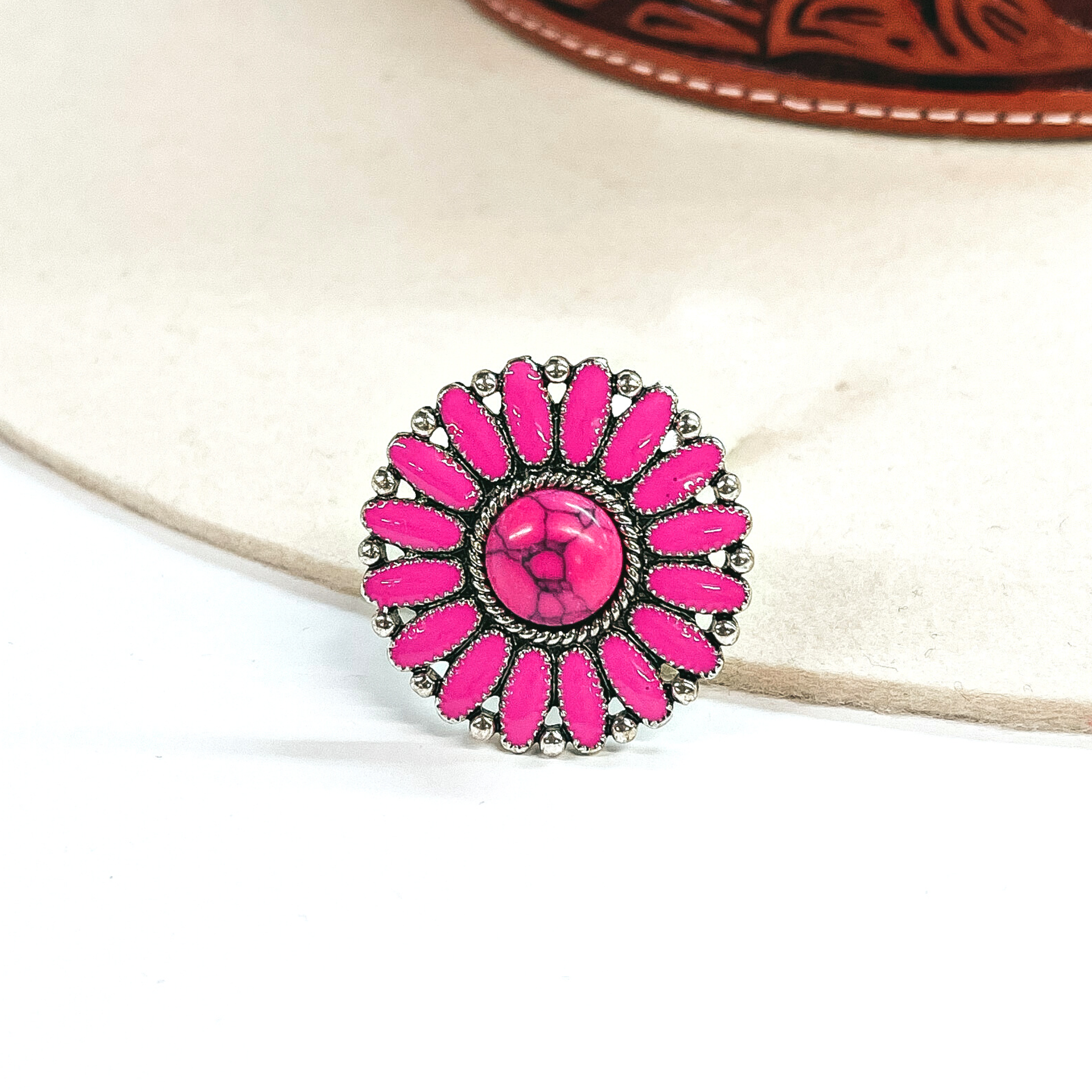 This is a pink,circle concho, epoxy cluster ring in a silver setting.  The ring has a small circle stone in the center with a silver, rope textured outline,  with oval shaped stone all around like a flower. In between each 'petal' it has silver  circles all around. This ring is taken on an ivory felt hat  with a brown textured band and on a white background.