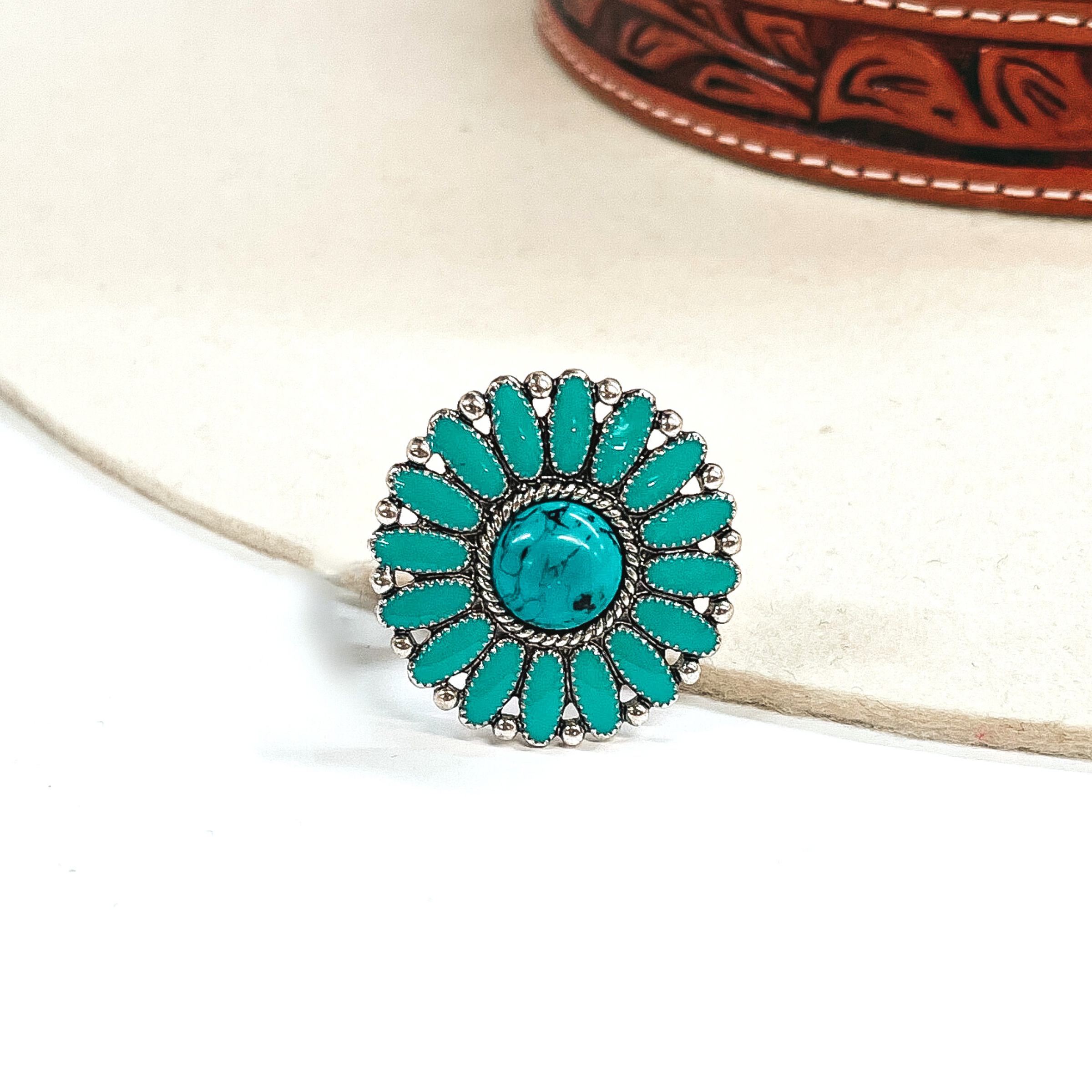 This is a turquoise,circle concho, epoxy cluster ring in a silver setting.  The ring has a small circle stone in the center with a silver, rope textured outline,  with oval shaped stone all around like a flower. In between each 'petal' it has silver  circles all around. This ring is taken on an ivory felt hat  with a brown textured band and on a white background.