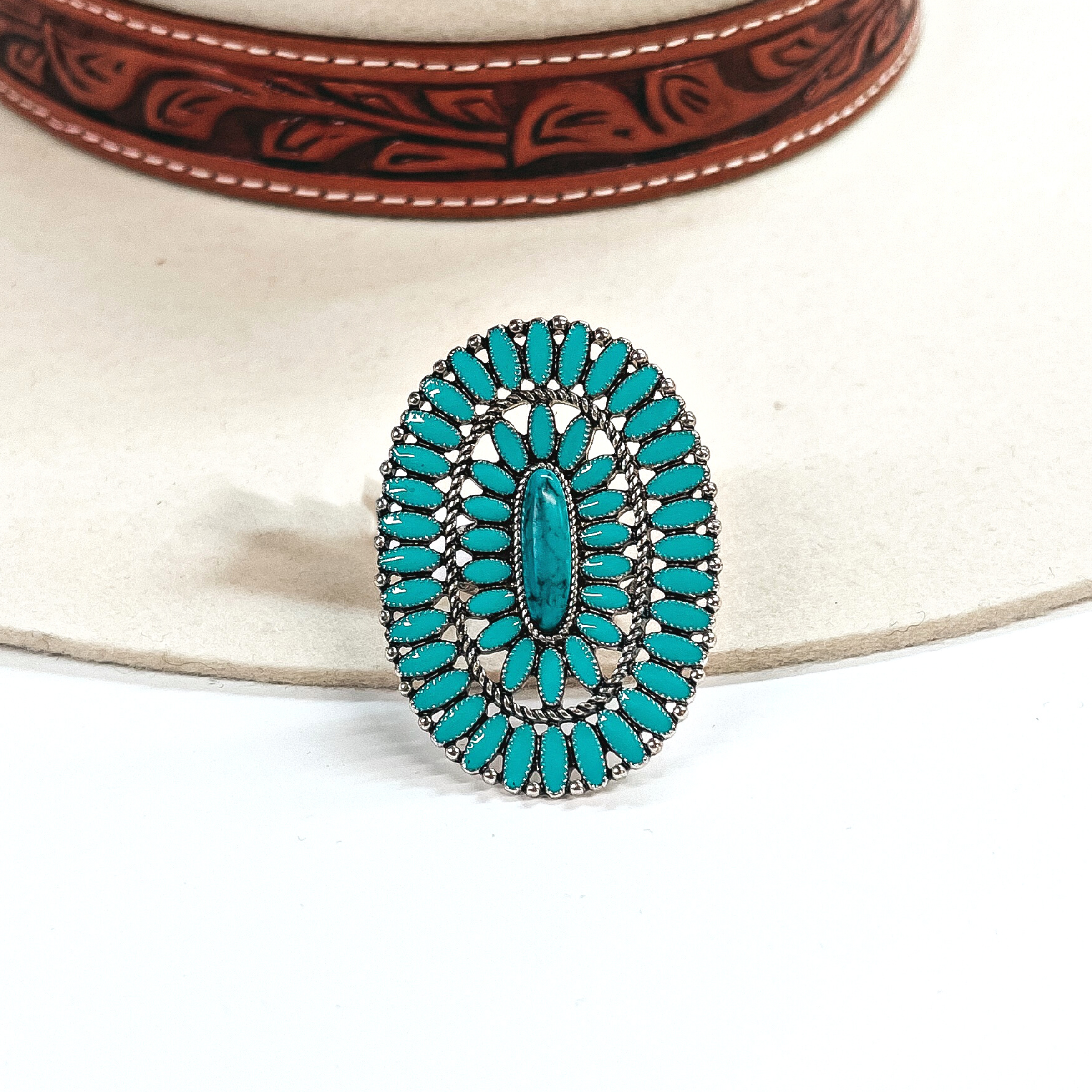 This is an oval turquoise stone cluster ring in a silver tone setting. There is a  long-skinny stone in the center with a silver textured outline with two rows of  small-oval stones all around. In between each row there is a silver rope-textured  outline. This ring is taken on an ivory felt hat  with a brown textured band and on a white background.