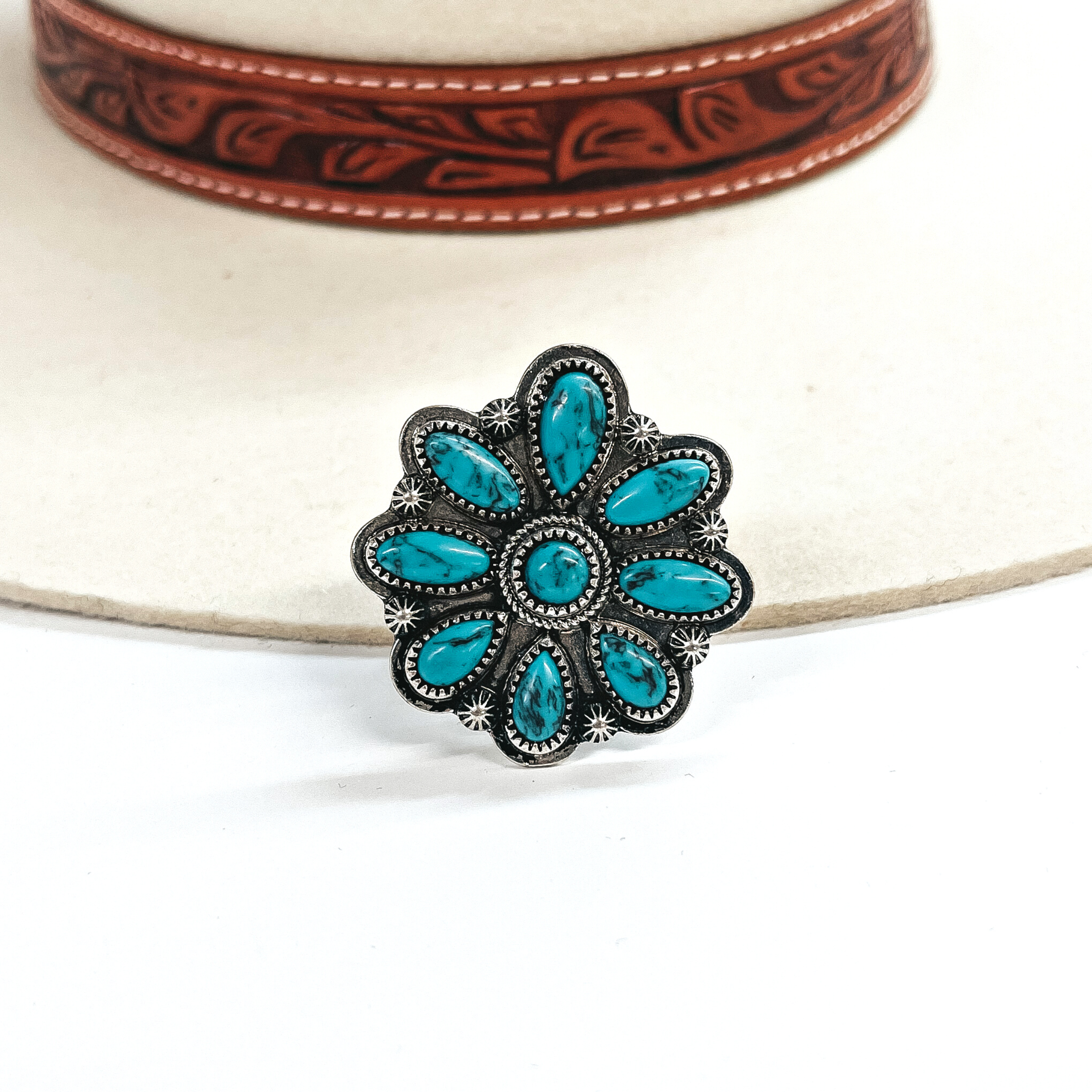 This a siver western flower with small turquoise stones in the petal setting.  This ring is taken on an ivory felt hat  with a brown textured band and on a white background.