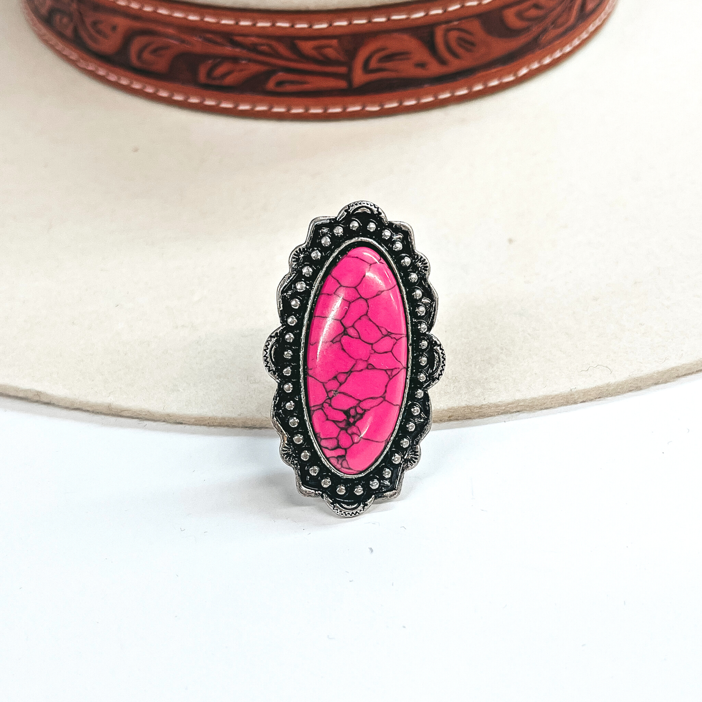 This is a long oval pink stone ring in a silver tone setting. The setting has  rounded bumps with small detailing all around. This ring is taken on an ivory felt hat  with a brown textured band and on a white background.