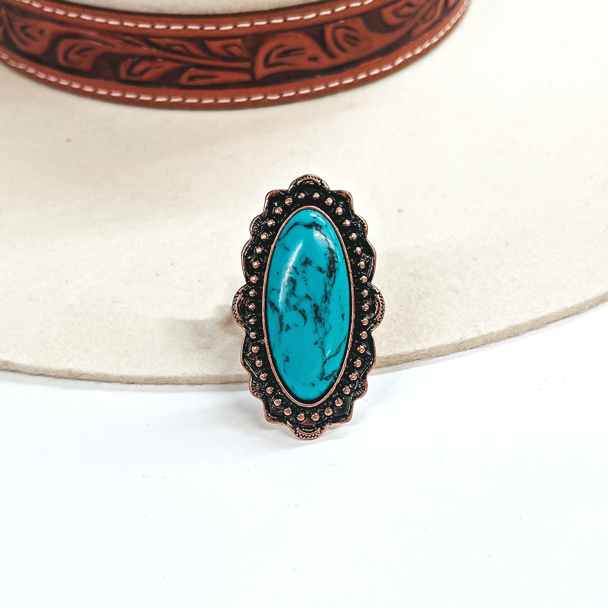 This is a long oval turquoise stone ring in a copper tone setting. The setting has  rounded bumps with small detailing all around. This ring is taken on an ivory felt hat  with a brown textured band and on a white background.