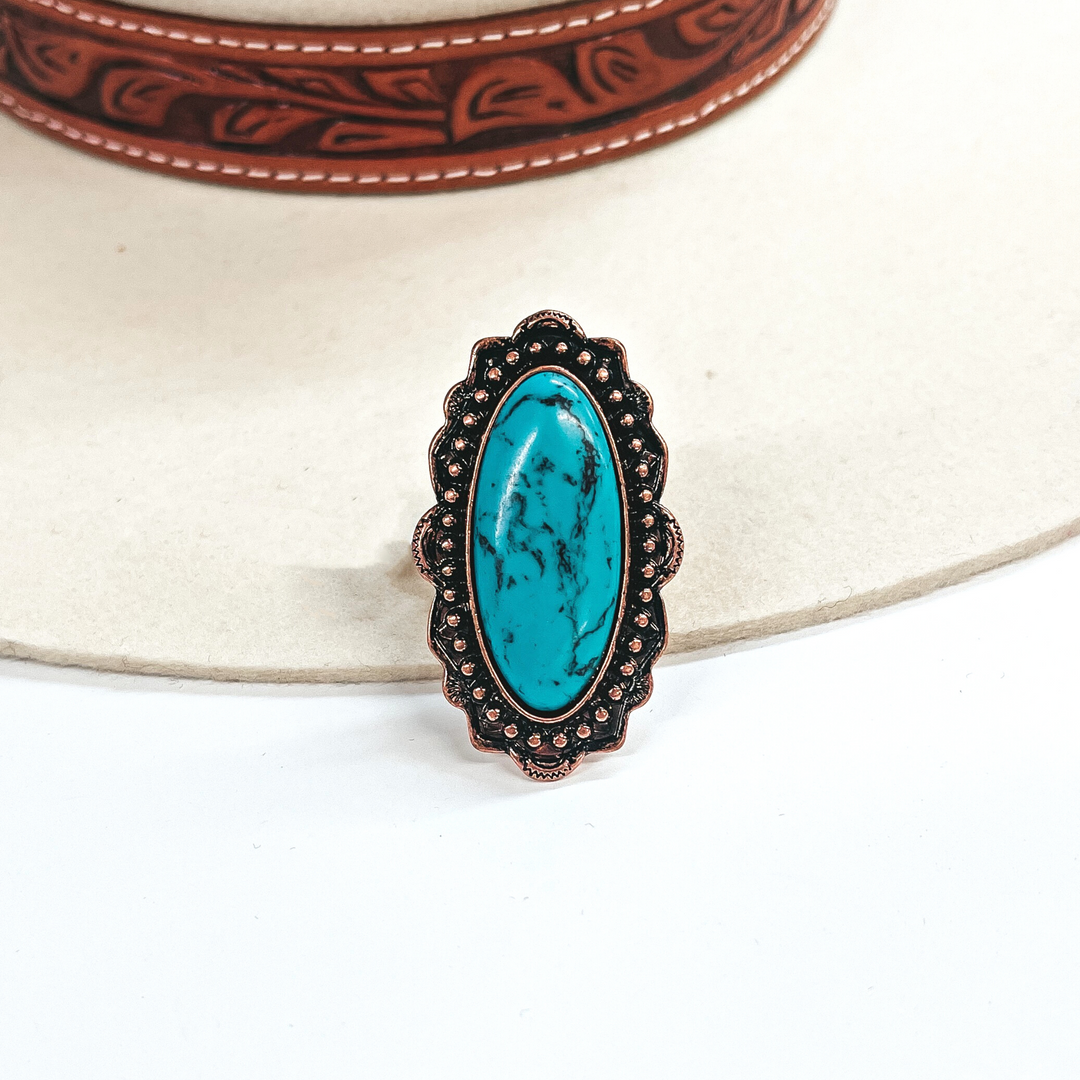 This is a long oval turquoise stone ring in a copper tone setting. The setting has  rounded bumps with small detailing all around. This ring is taken on an ivory felt hat  with a brown textured band and on a white background.