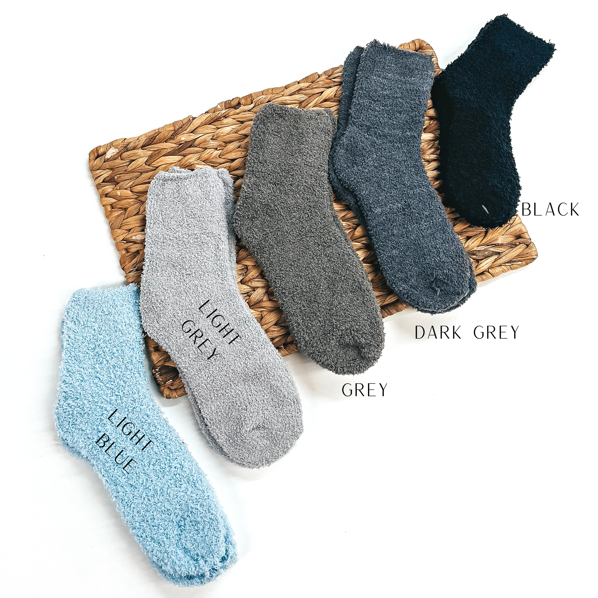 These are five pairs of fuzzy socks, from left to right; light blue, light grey, grey,  dark grey, and black. These socks are taken on a brown woven slate and on a white  background.
