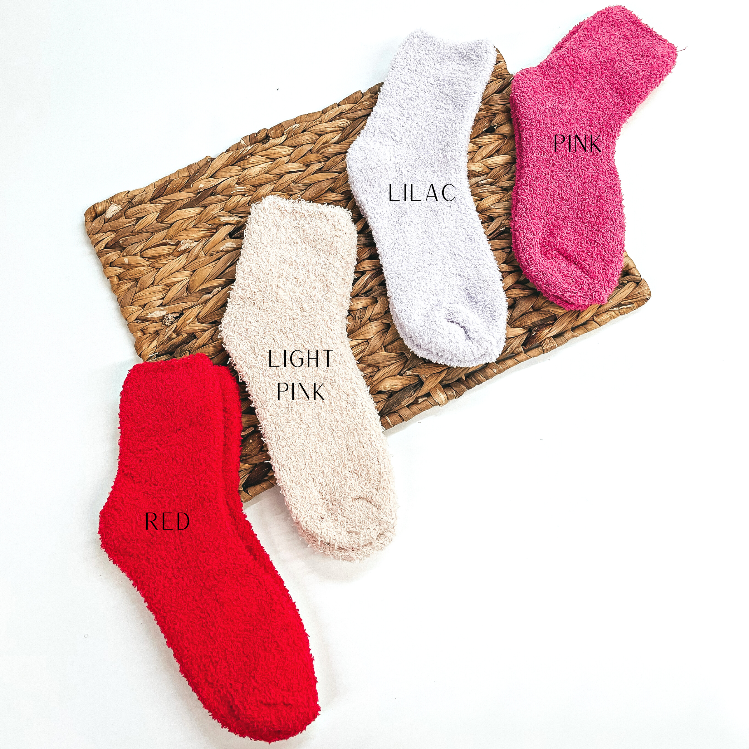 These are four pairs of fuzzy socks, from left to right; red, light pink, lilac,  and pink. These socks are taken on a brown woven slate and on a white  background.