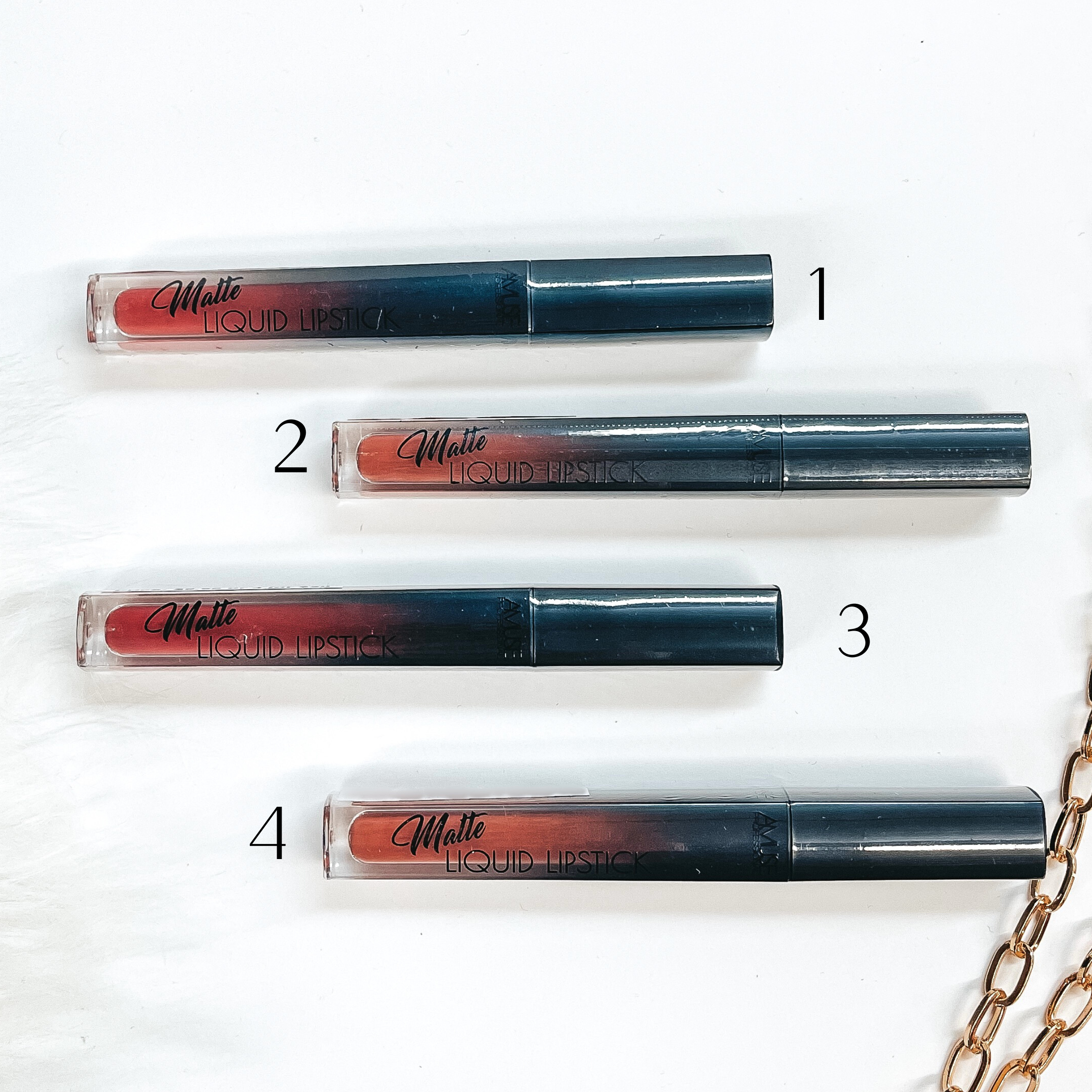 There are four liquid lipsticks in different shades of pink-nudes laying on a white  background with white fur and a gold link chain in the side as decor.