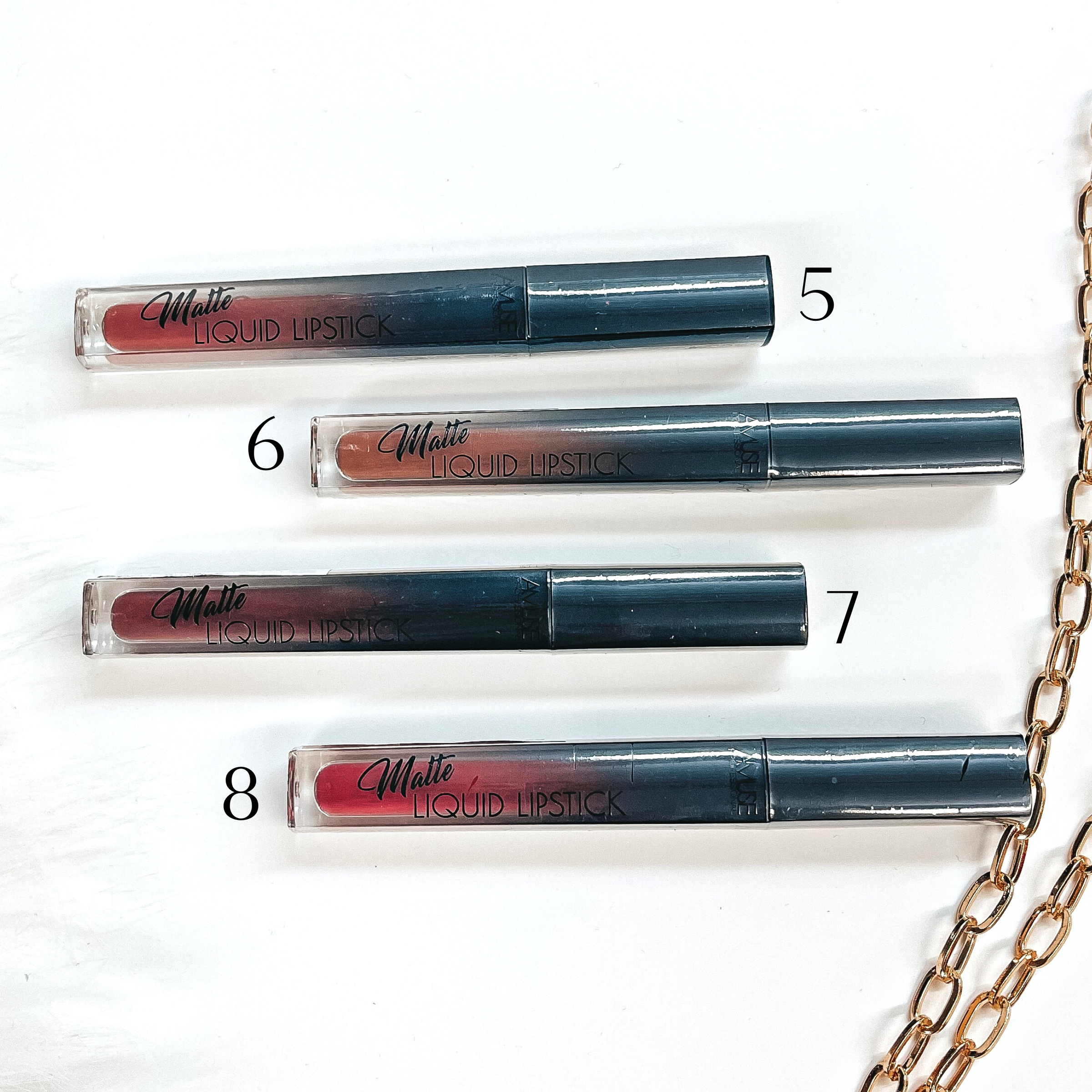 There are four liquid lipsticks in different shades of dark-nudes/red laying on a white  background with white fur and a gold link chain in the side as decor.
