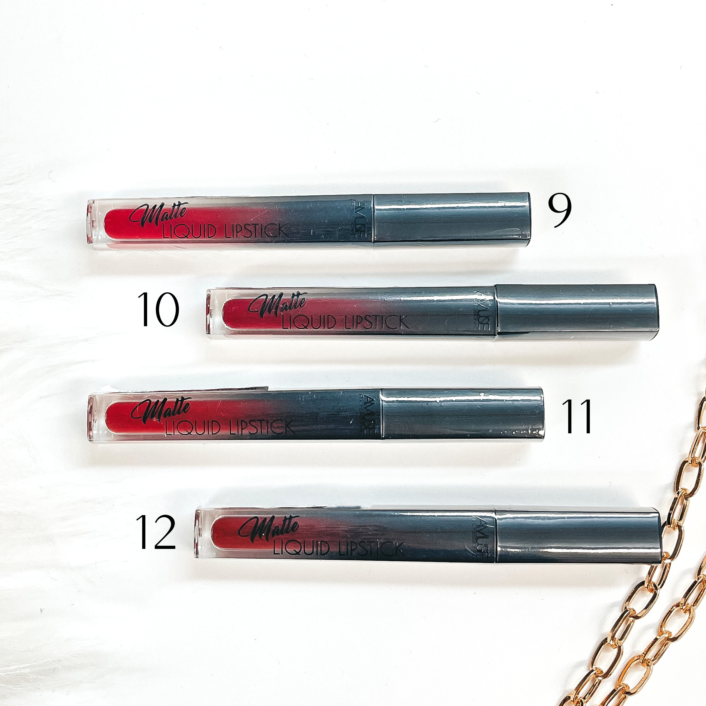 There are four liquid lipsticks in different shades of dark/bright reds laying on a white  background with white fur and a gold link chain in the side as decor.