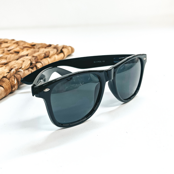 This is a black frame round sunglasses with a black,dark grey lense with small silver  detailing. This pair of sunglasses are taken on a white background with a brown  woven slate in the side as decor.