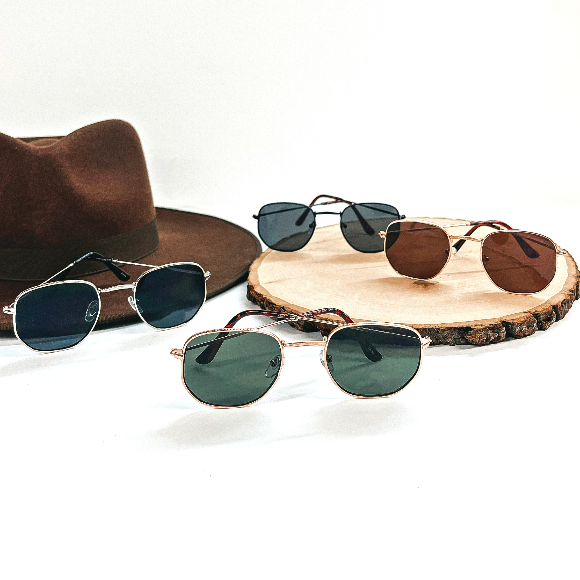 These are four pairs of sunglasses in silver, black, gold and rose gold frame/outline. These sunglasses are taken on a wooden slate, white background, and a brown felt hat  in the back as decor.