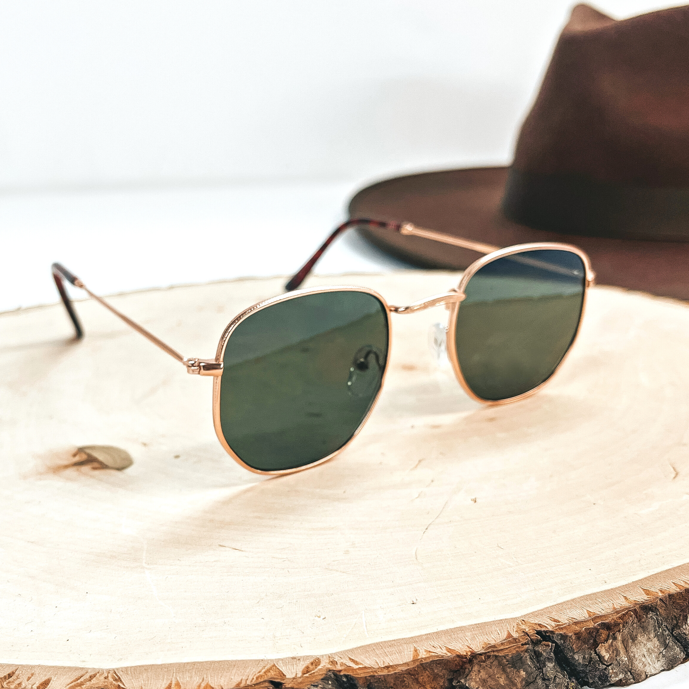This is a rose gold outlined pair of sunglasses with a dark green lense with  tortouise print ear pieces. This pair of sunglasses on a wooden slate with a dark brown  felt hat in the back as decor.