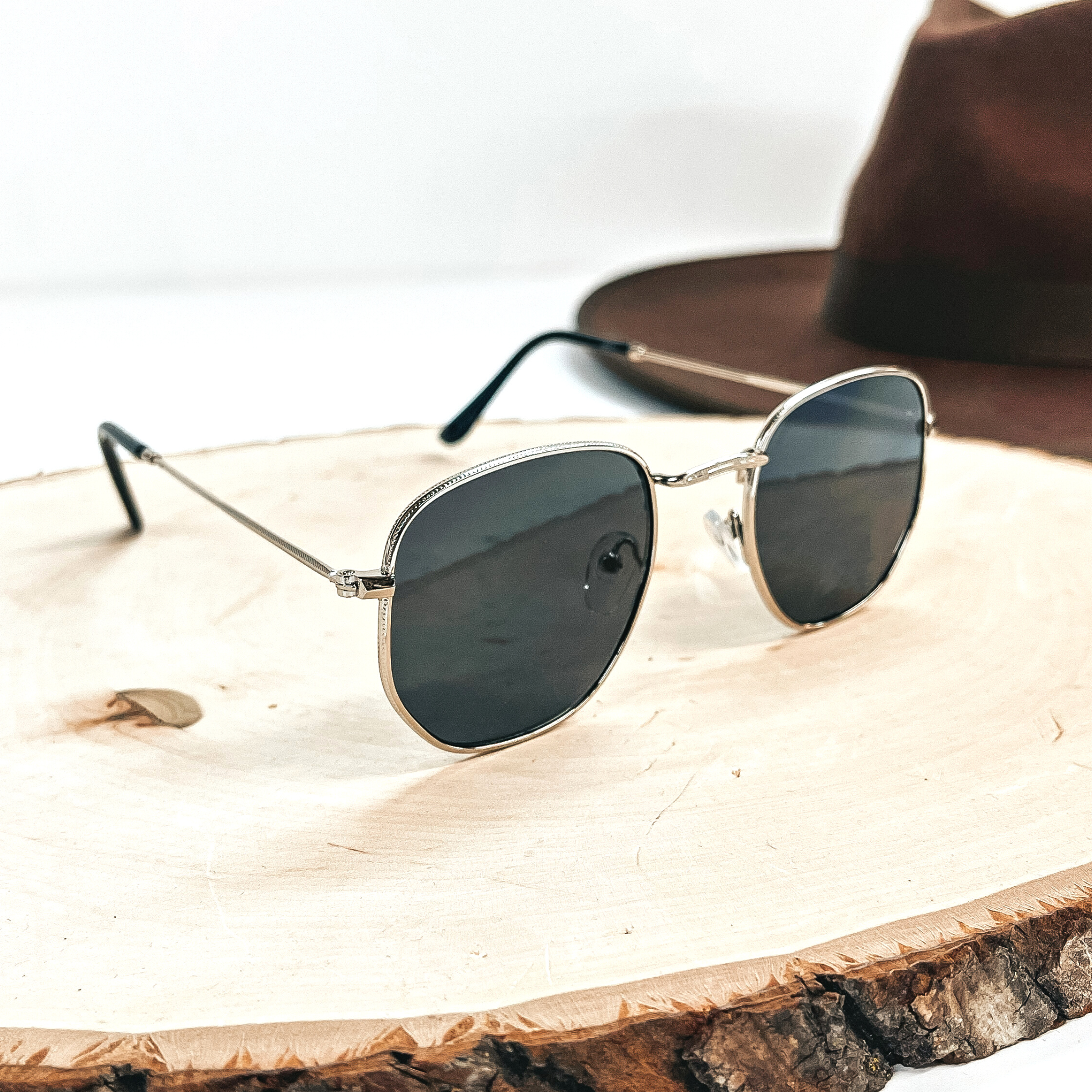 This is a silver outlined pair of sunglasses with a black/dark grey lense with  black ear pieces. This pair of sunglasses on a wooden slate with a dark brown  felt hat in the back as decor.