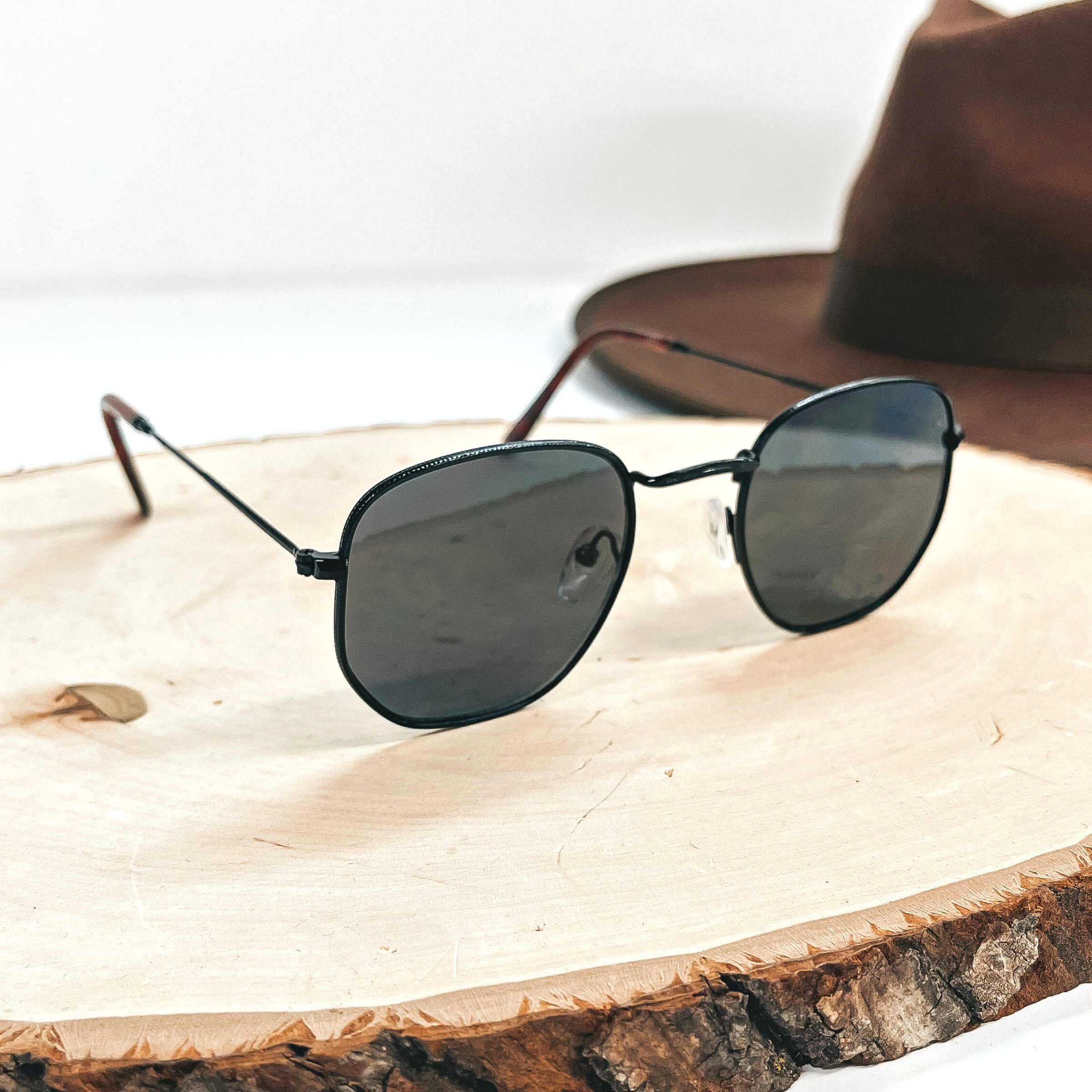 This is a black outlined pair of sunglasses with a black/dark grey lense with  brown ear pieces. This pair of sunglasses on a wooden slate with a dark brown  felt hat in the back as decor.