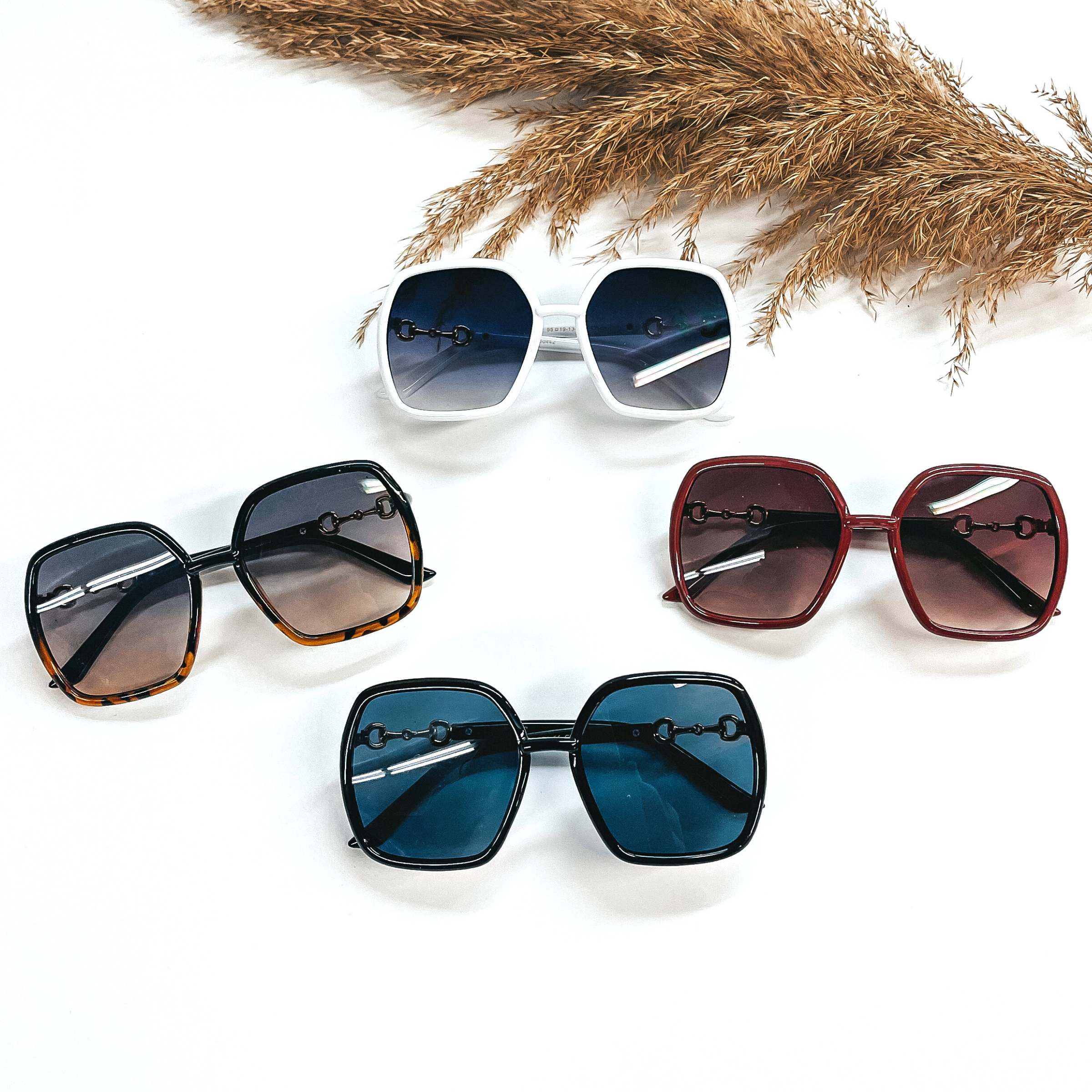 These are four pairs of big square sunglasses in white, tortouise, maroon, and black. They are laying on white background with a brown plant on the top as decor.