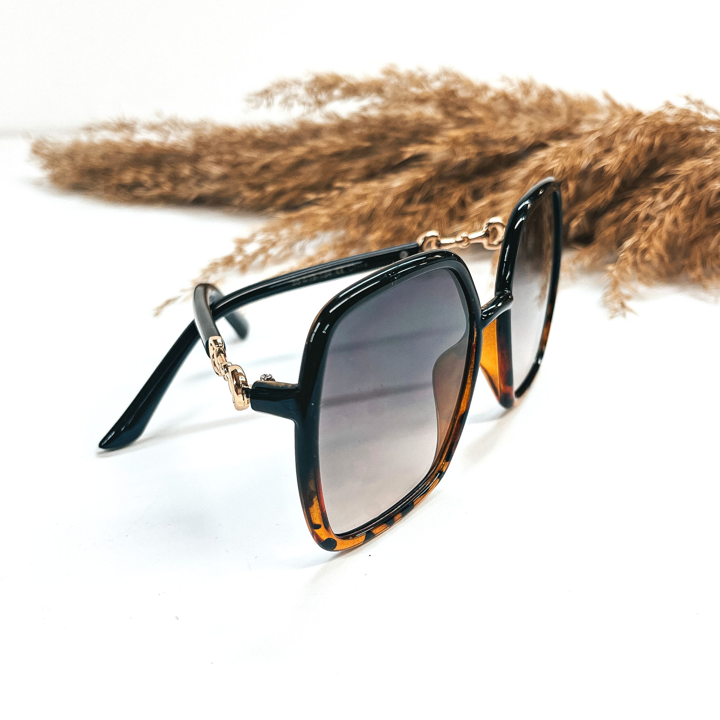 This is a pair of black and tortuoise print frame, large, square sunglasses with a  black/dark grey lense.  The side of the glasses have gold detailing as a connector. These pair of  sunglasses are taken on a white background with a brown plant in the back as decor.