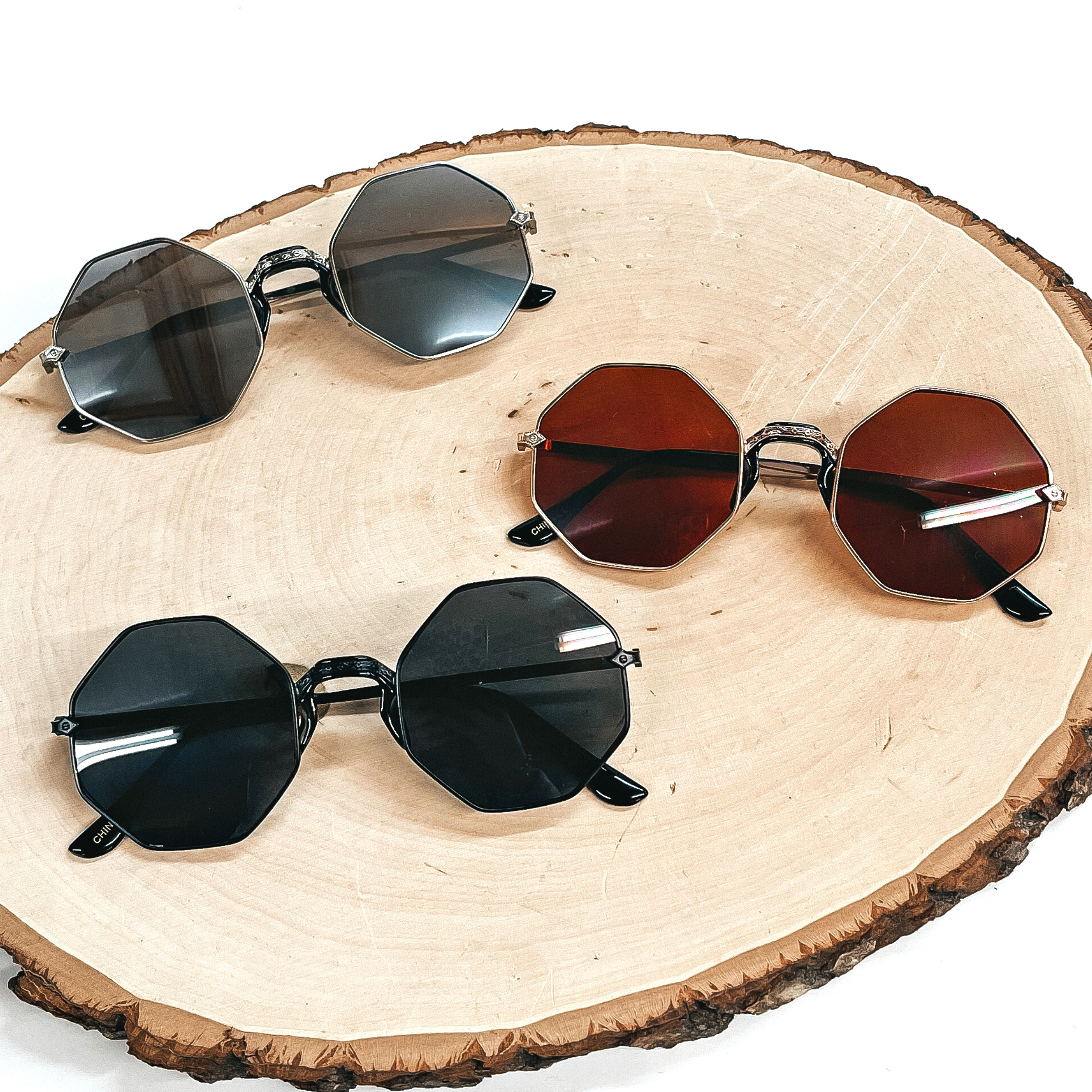 These are three pairs of octagon sunglasses in black, brown, and silver. These sunglasses are taken on a  brown wooden slate and on a white background.