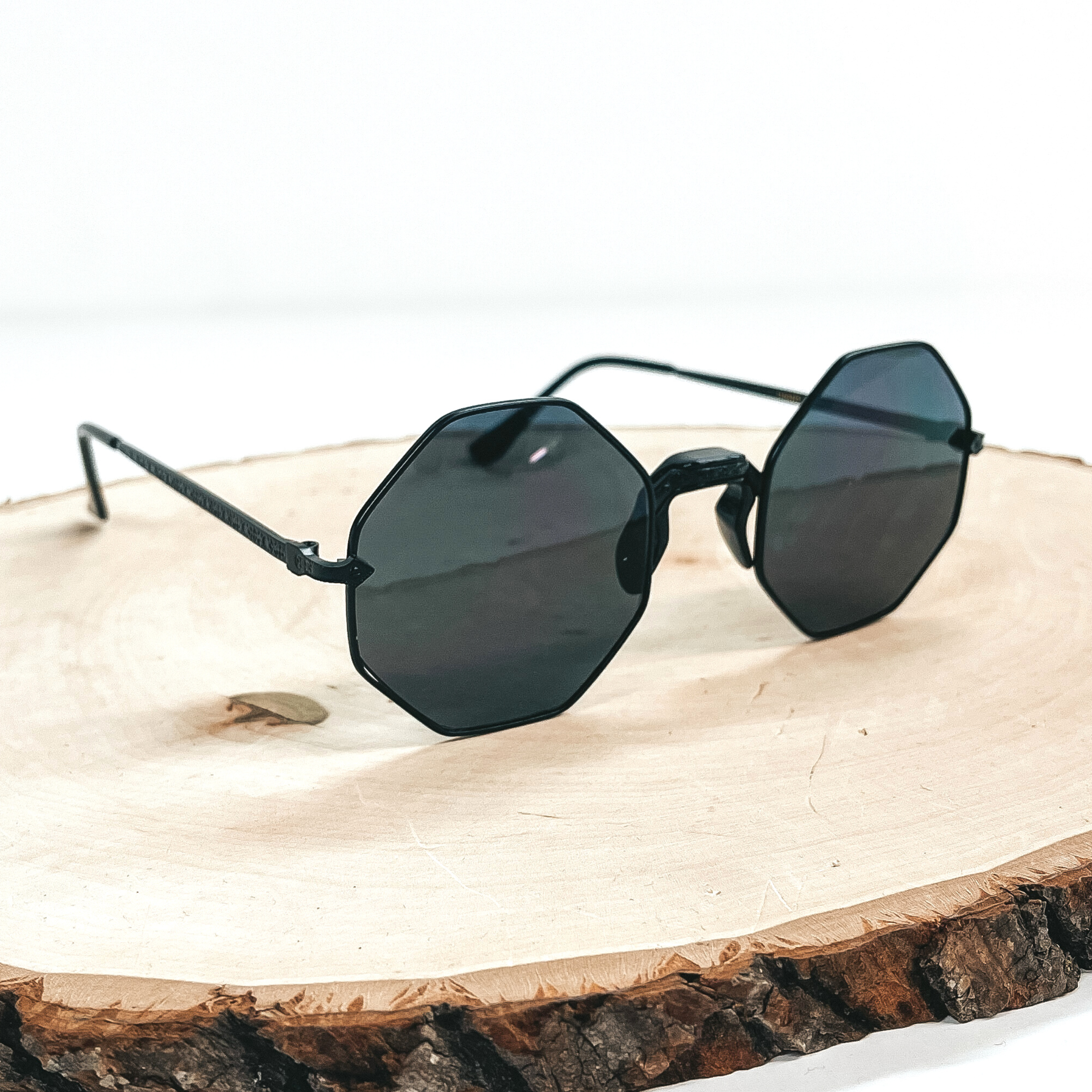 This is a pair of black octagon sunglasses, the lense is black/dark grey with a  black outline. These pair of sunglasses are taken on a wooden slate and on a white  background.