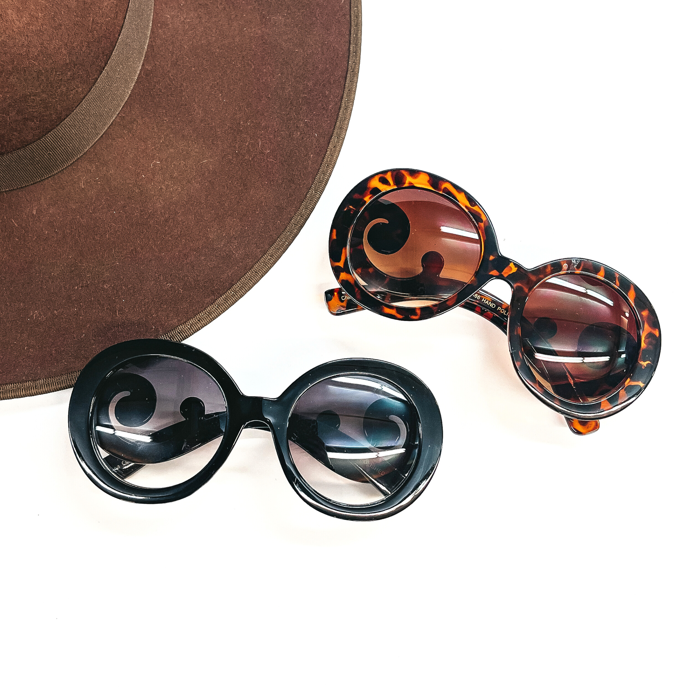 These are two pairs of round sunglasses in different colors/prints. One pair is  black frame with a black/dark grey lense. The second pair is tortuoise print frame with a  brown lense. These sunglasses are taken laying on a white background and a dark brown  felt hat in the side as decor.