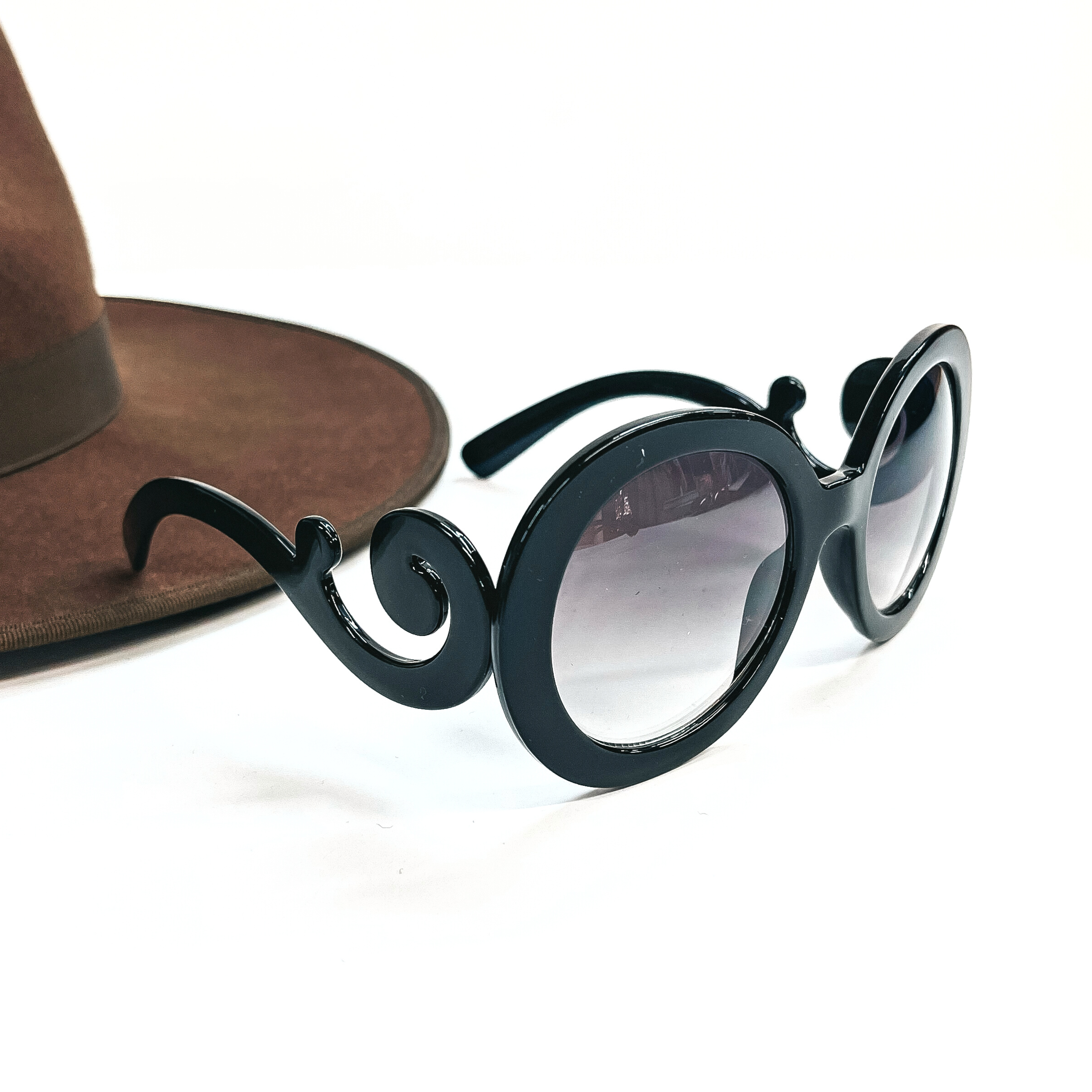 This is a black pair of sunglasses, the frame is black with a black/dark grey lense. The side parts of the sunglasses have a swirl. These sunglasses are taken on a white  background with a dark brown felt hat in the side as decor.