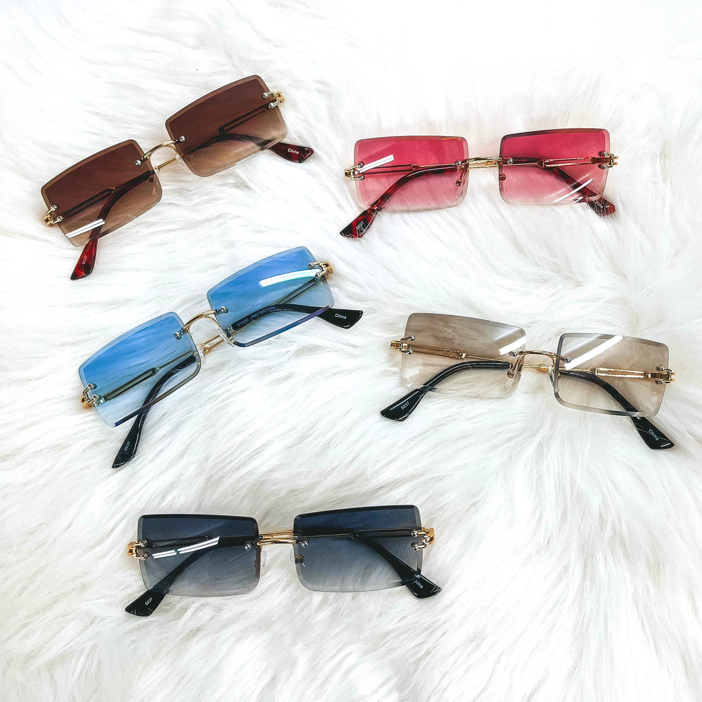 There are five pairs of colorful sunglasses in brown, pink, blue, clear brown/grey,  and black/dark grey. All sunglasses have no frame except gold connectors. These  sunglasses are taken laying on a white fur.