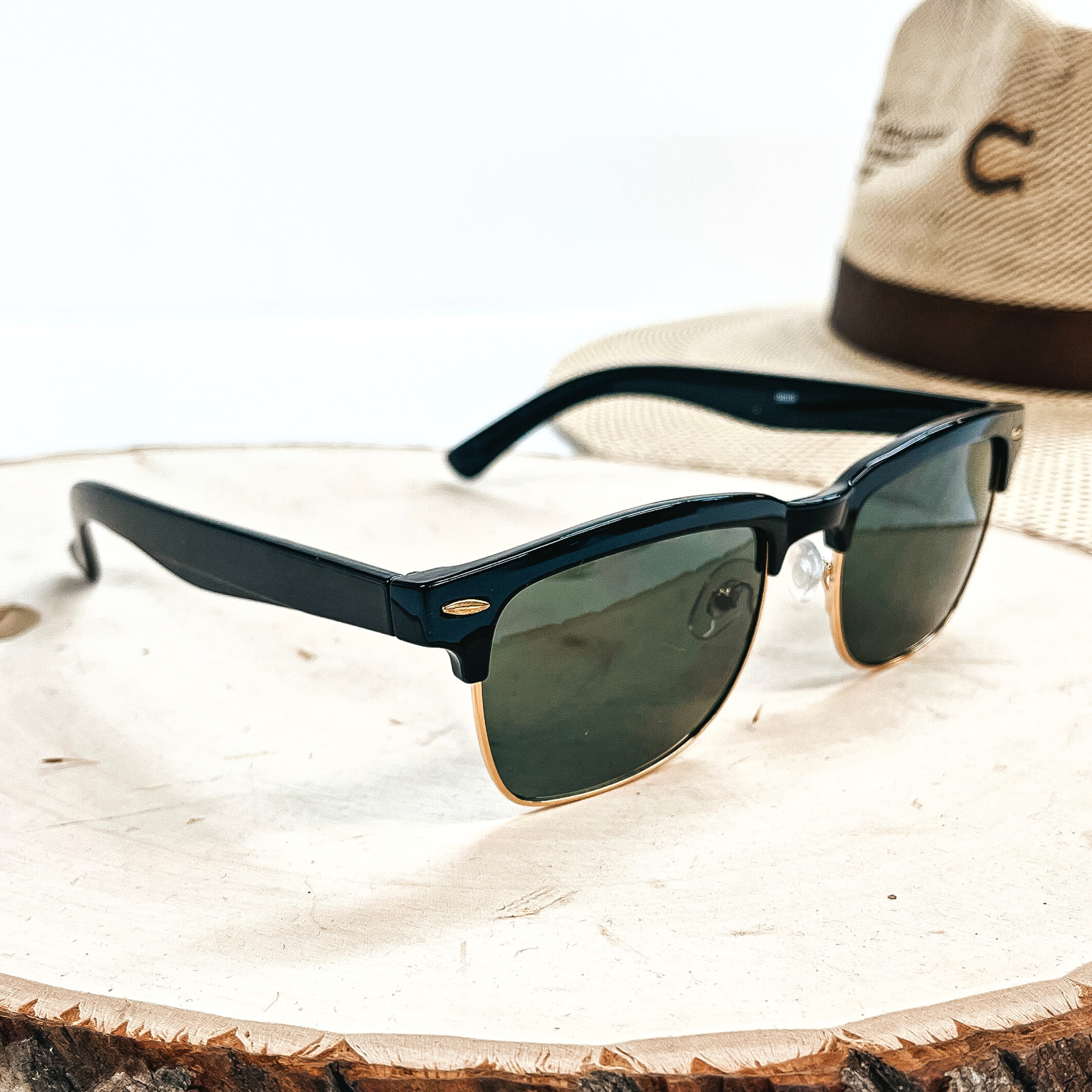This is a pair of black sunglasses with a black/dark grey lense and a gold  outline. These sunglasses are taken on top of a wooden slate with a straw hat in the  back as decor.