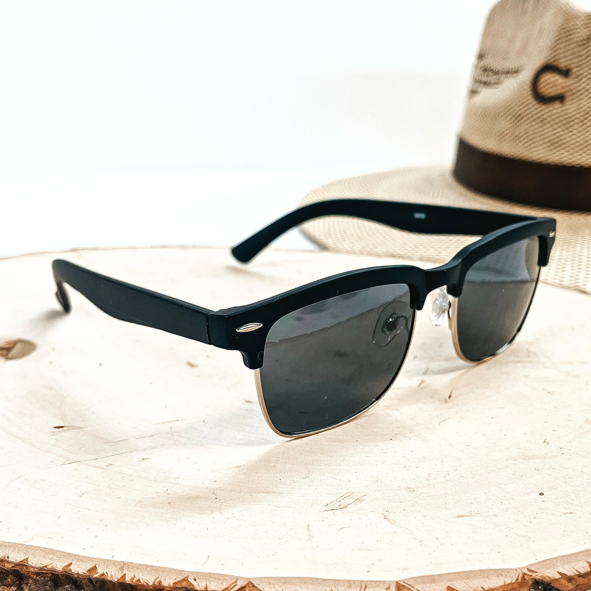 This is a pair of black matte sunglasses with a dark grey/black  lense and a silver  outline. These sunglasses are taken on top of a wooden slate with a straw hat in the  back as decor.
