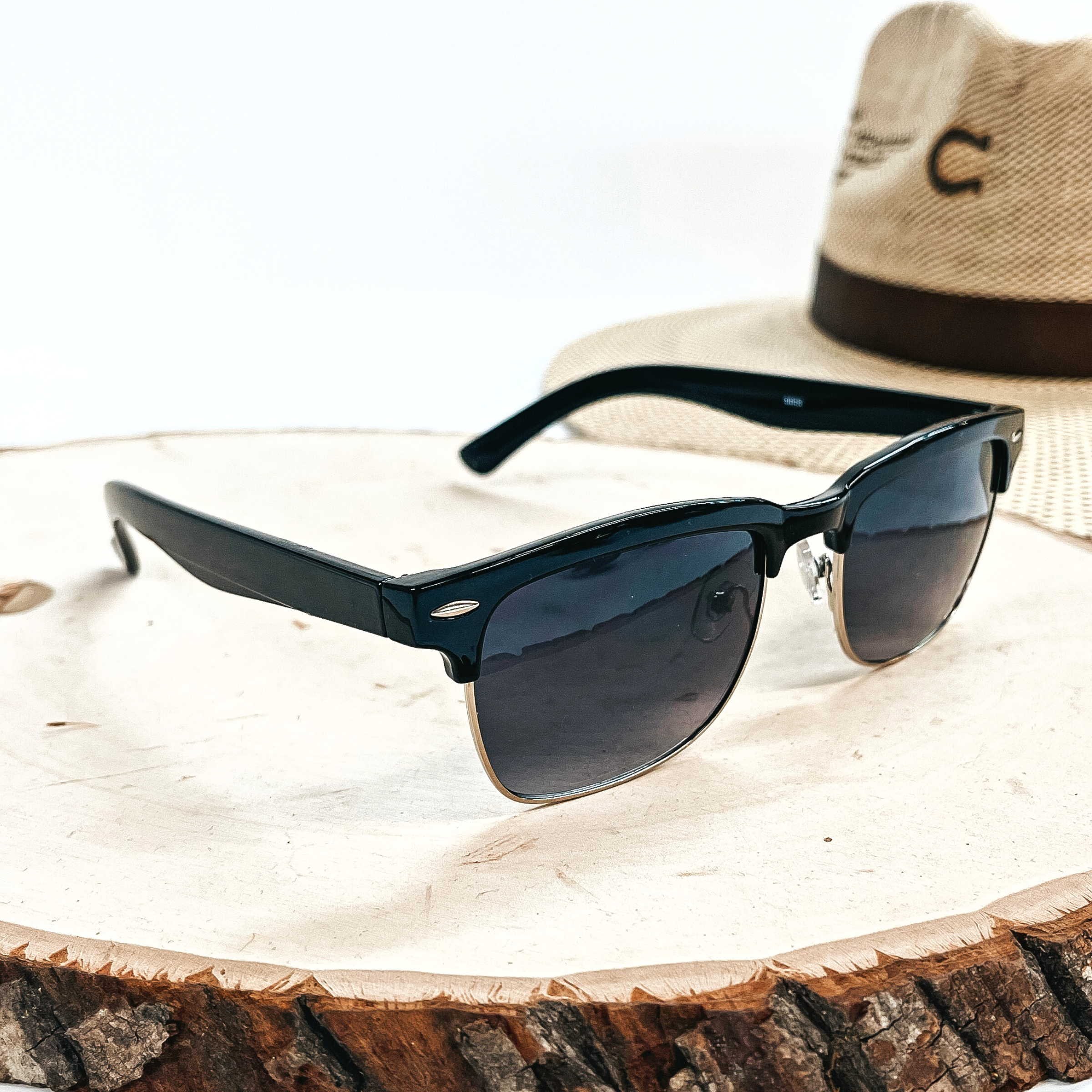 This is a pair of black sunglasses with a black/dark grey lense and a silver  outline. These sunglasses are taken on top of a wooden slate with a straw hat in the  back as decor.
