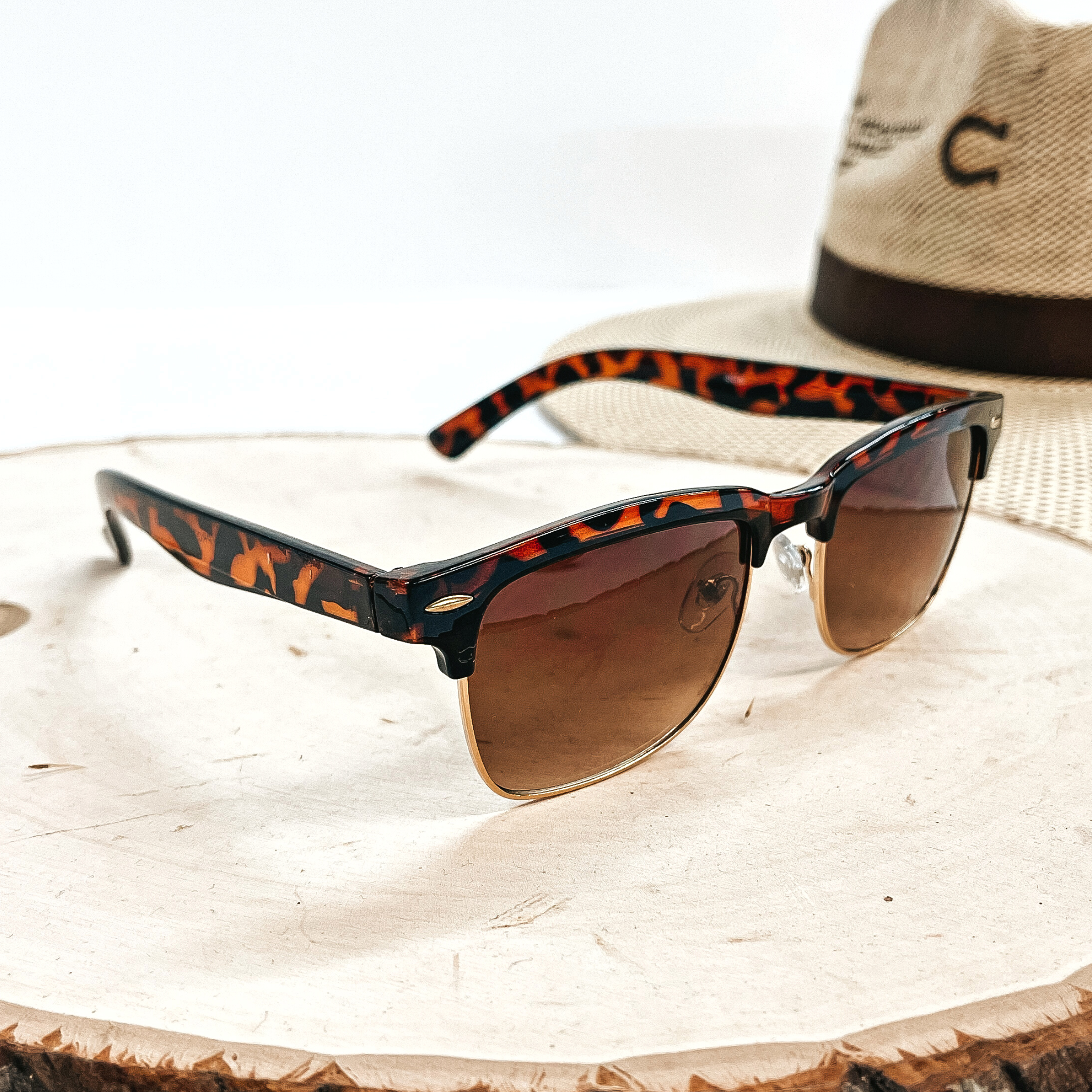 This is a pair of brown tortouise print sunglasses with a brown lense and a gold  outline. These sunglasses are taken on top of a wooden slate with a straw hat in the  back as decor.