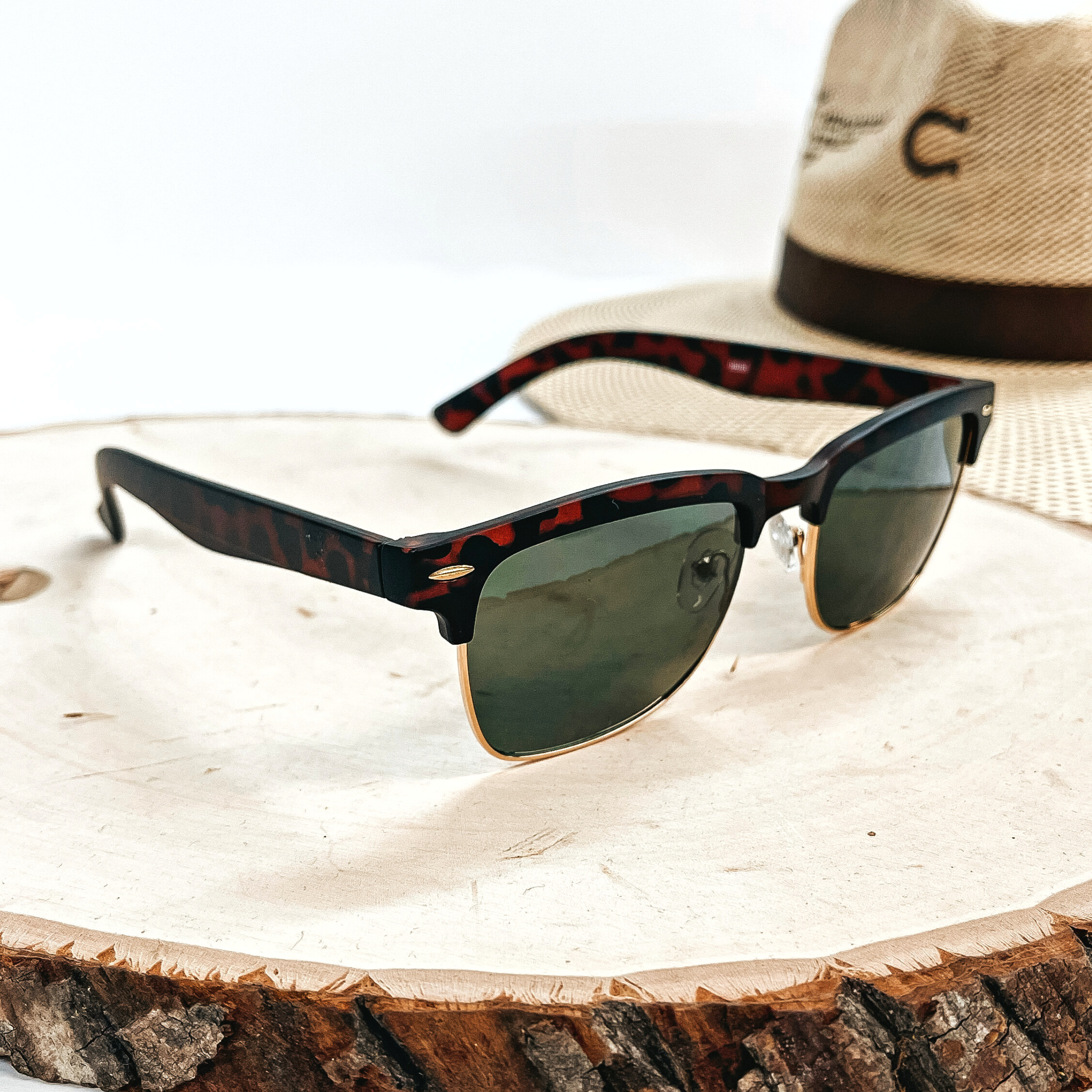 This is a pair of brown tortouise print matte sunglasses with a dark green  lense and a gold  outline. These sunglasses are taken on top of a wooden slate with a straw hat in the  back as decor.